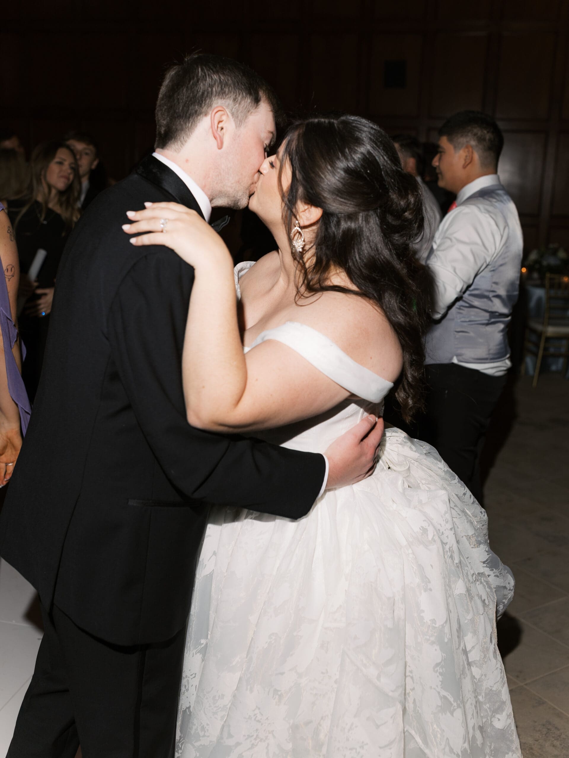 Bride and groom stealing a kiss on the dance floor during their wedding reception, surrounded by guests celebrating in the background.
