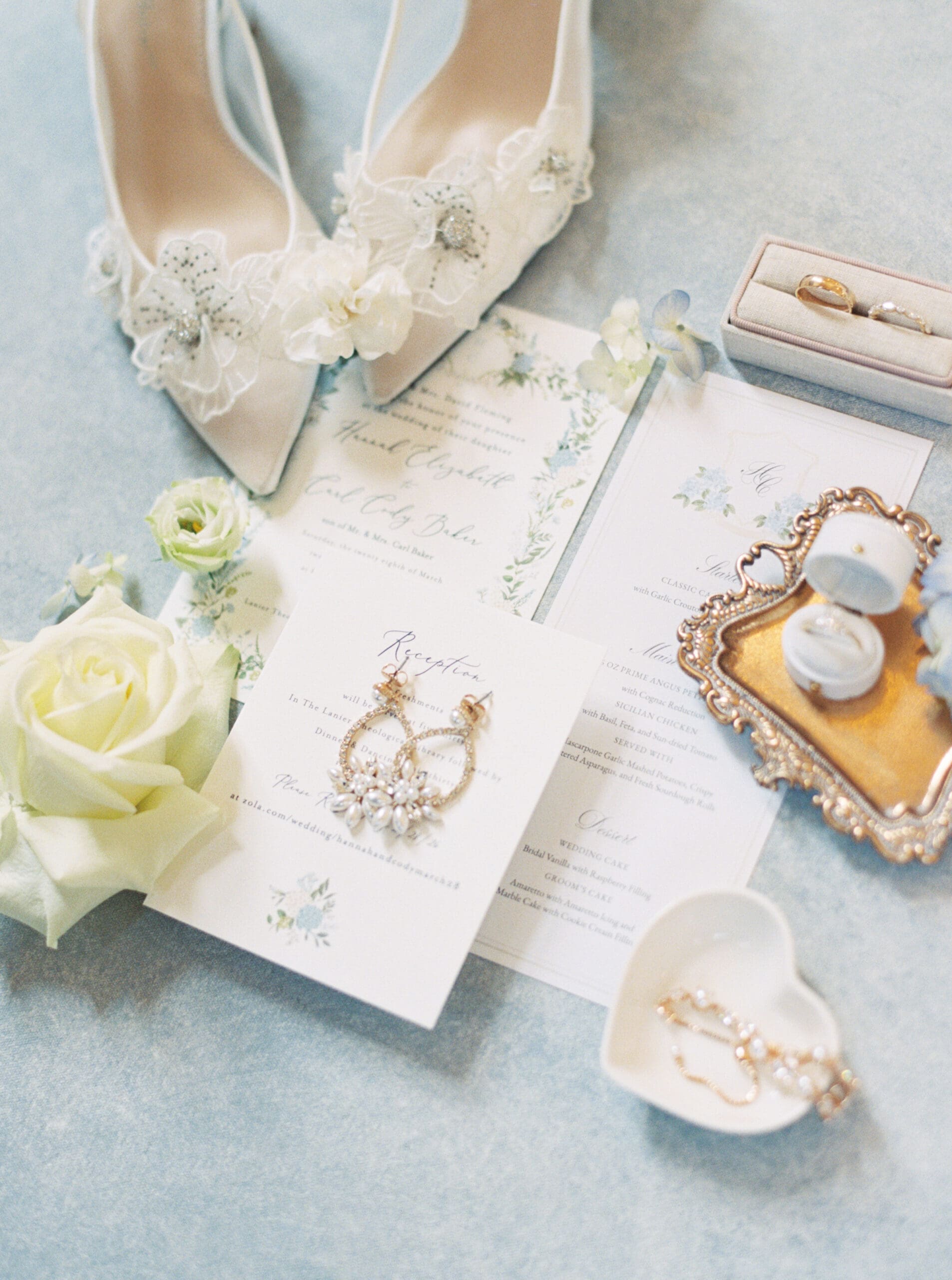 Flat lay of bridal details including embellished white heels, floral stationery, gold earrings, and wedding rings styled on a dusty blue surface.
