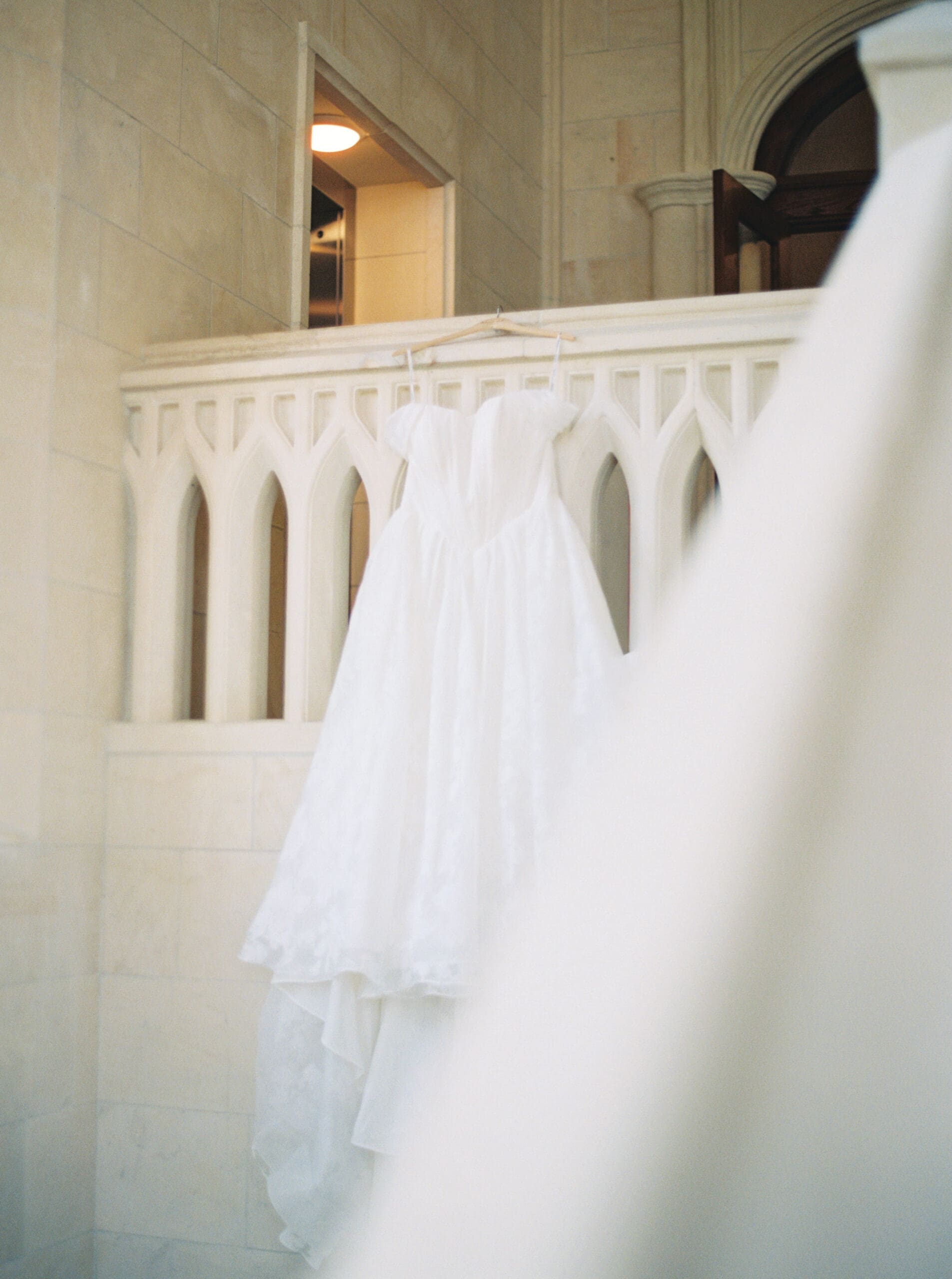 A white ball gown wedding dress hanging from an ornate Gothic stone balcony railing.
