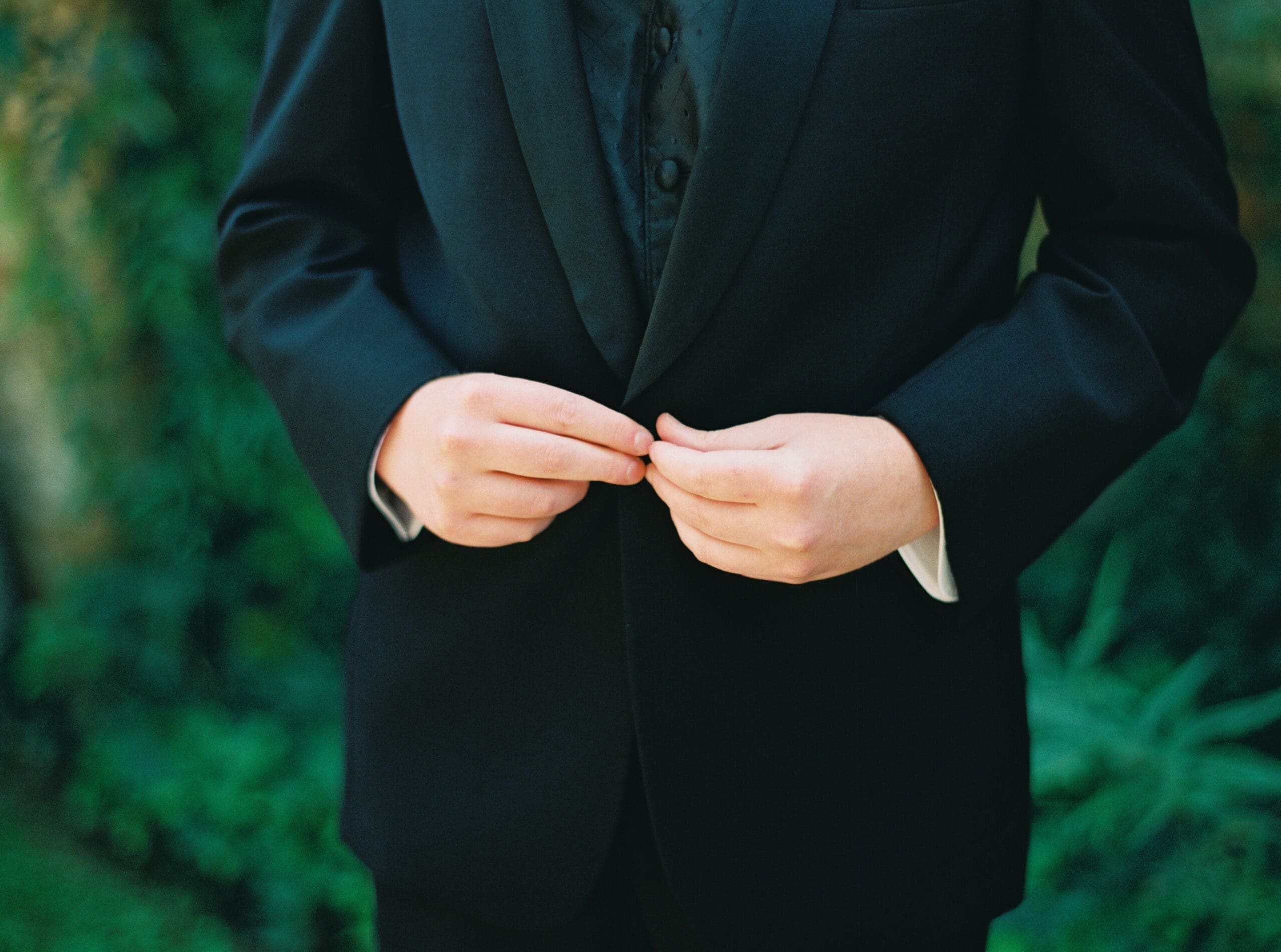 Close-up of a groom buttoning his black tuxedo jacket against a lush green garden backdrop.
