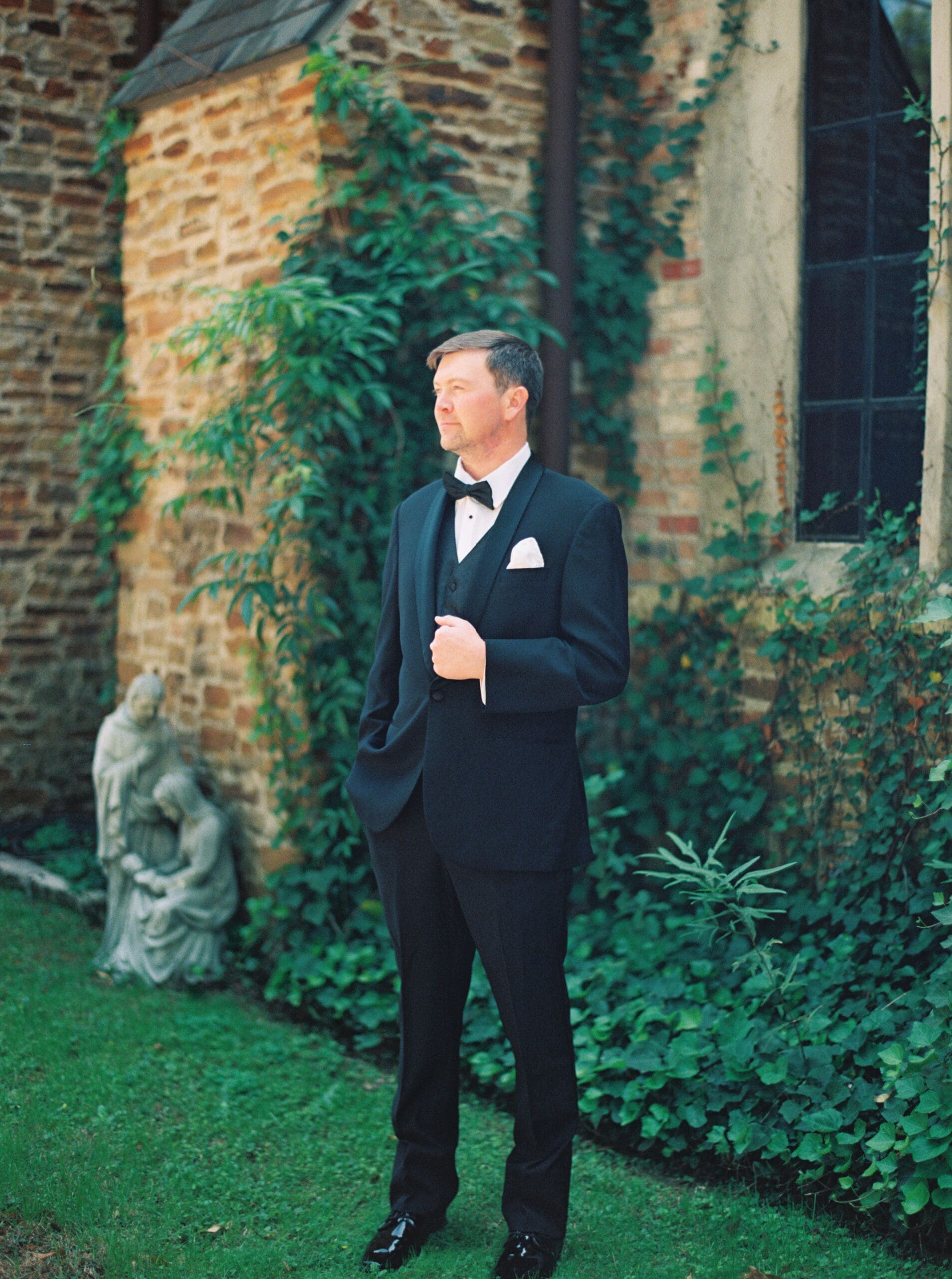 Groom standing in a classic black tuxedo beside the ivy-draped stone exterior of the wedding venue, gazing off into the distance with a stone garden statue beside him.
