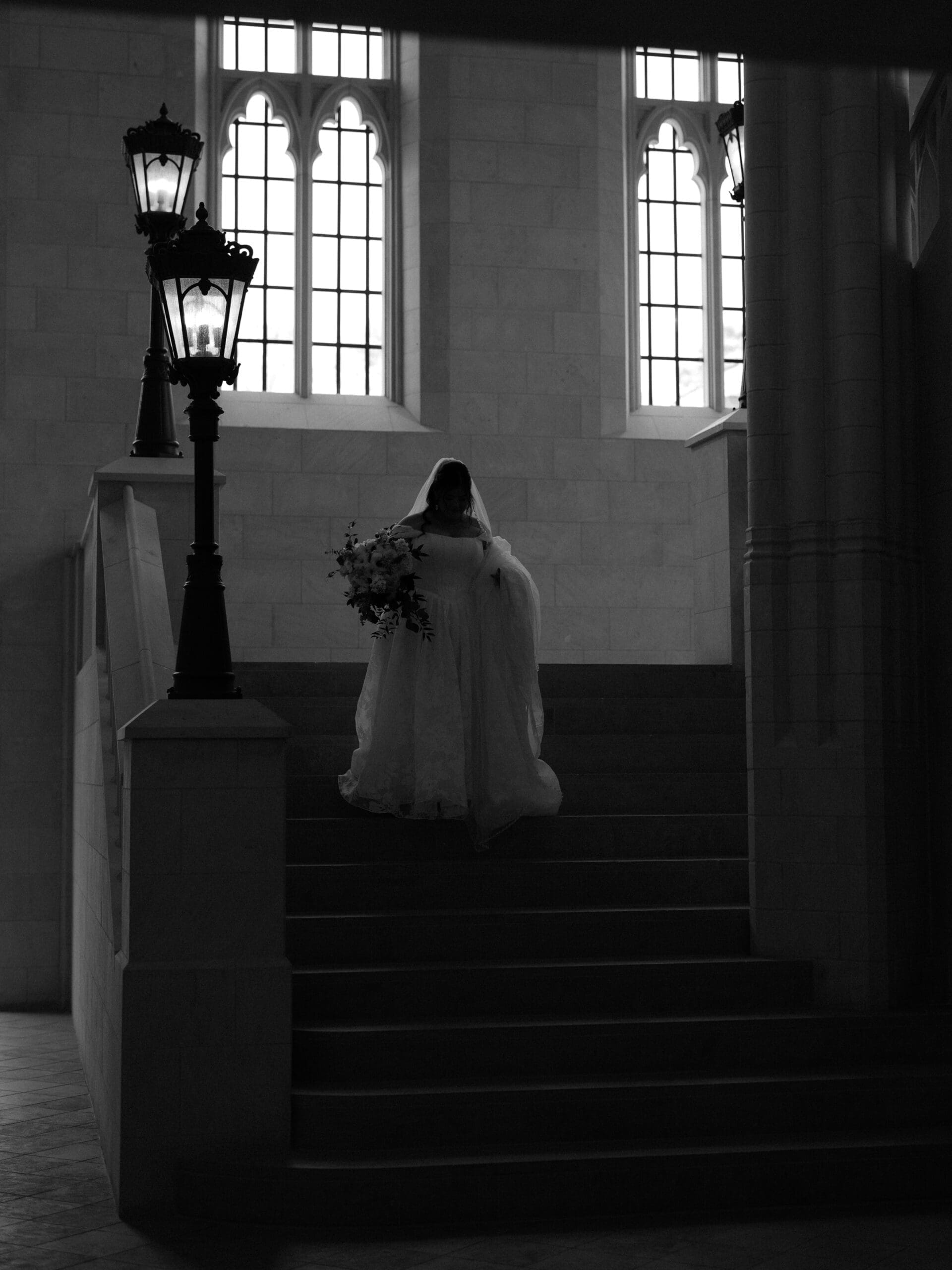 Black and white silhouette of a bride ascending the grand staircase at The Lanier Theological Library, holding her bouquet beneath arched Gothic windows.
