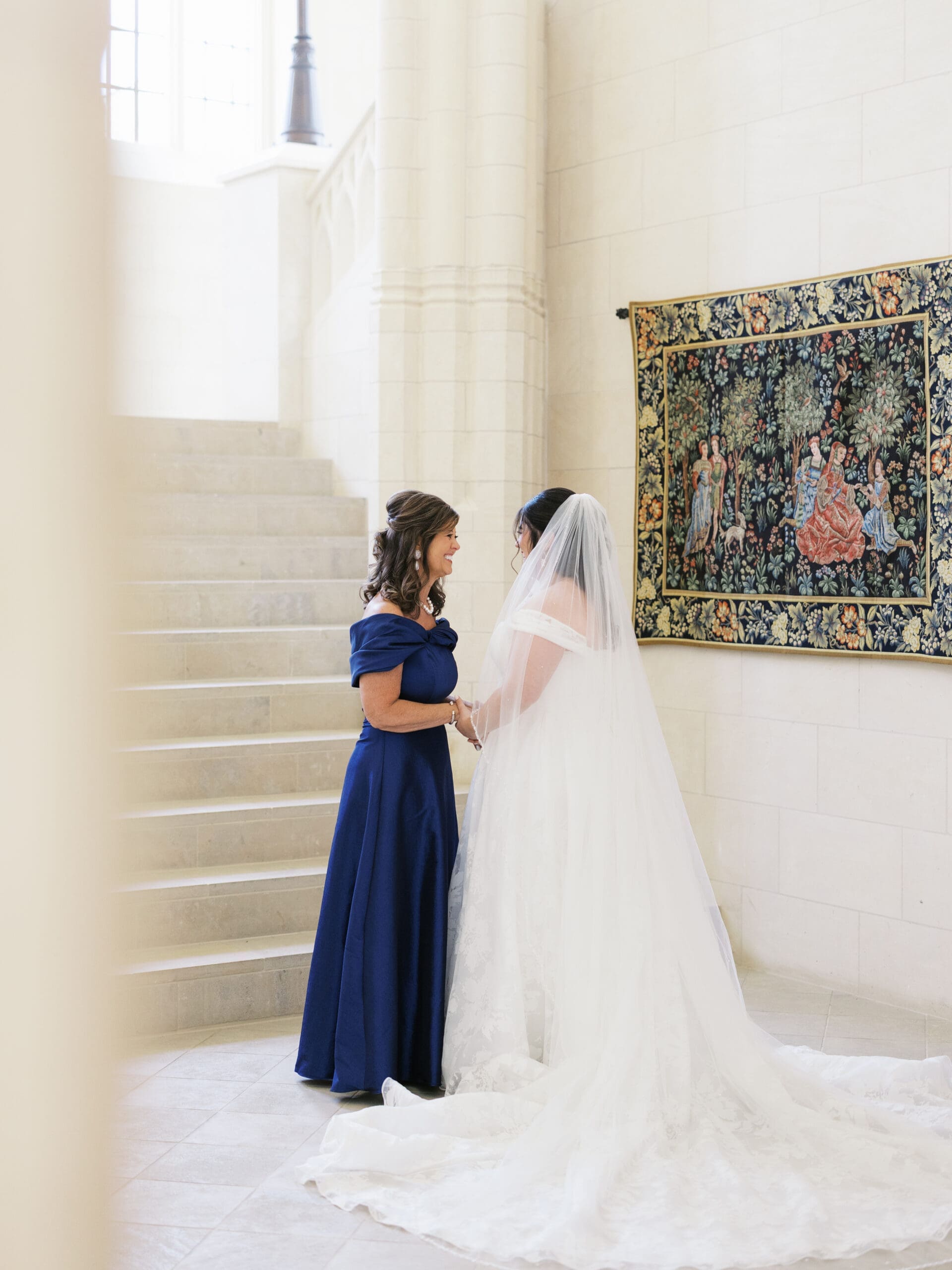 Bride and her mother sharing a tender moment on the marble staircase at The Lanier Theological Library, with a colorful tapestry on the wall behind them.
