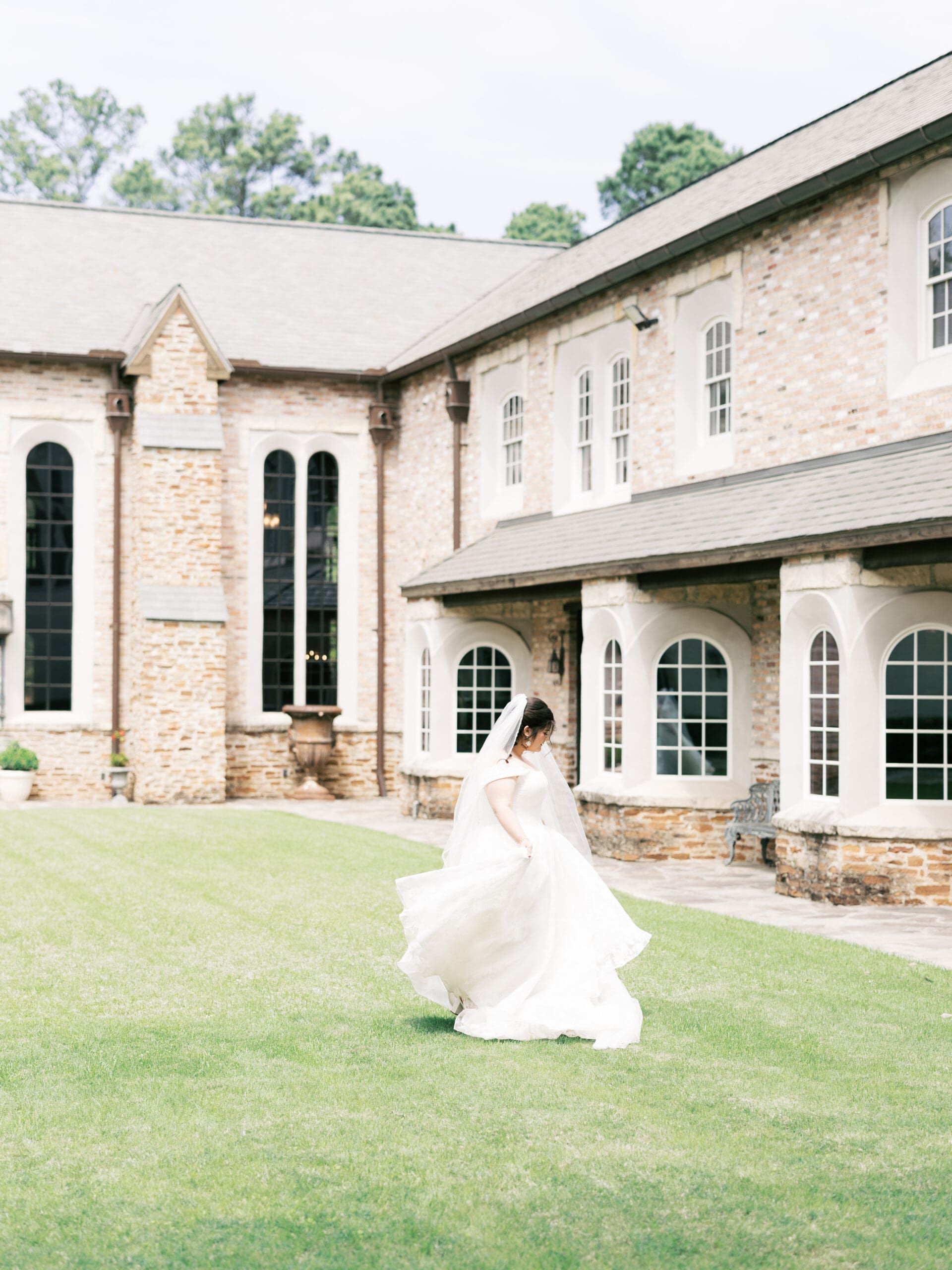 Bride twirling her lace ball gown on the green lawn beside The Lanier Theological Library, the stone exterior and arched windows visible behind her.
