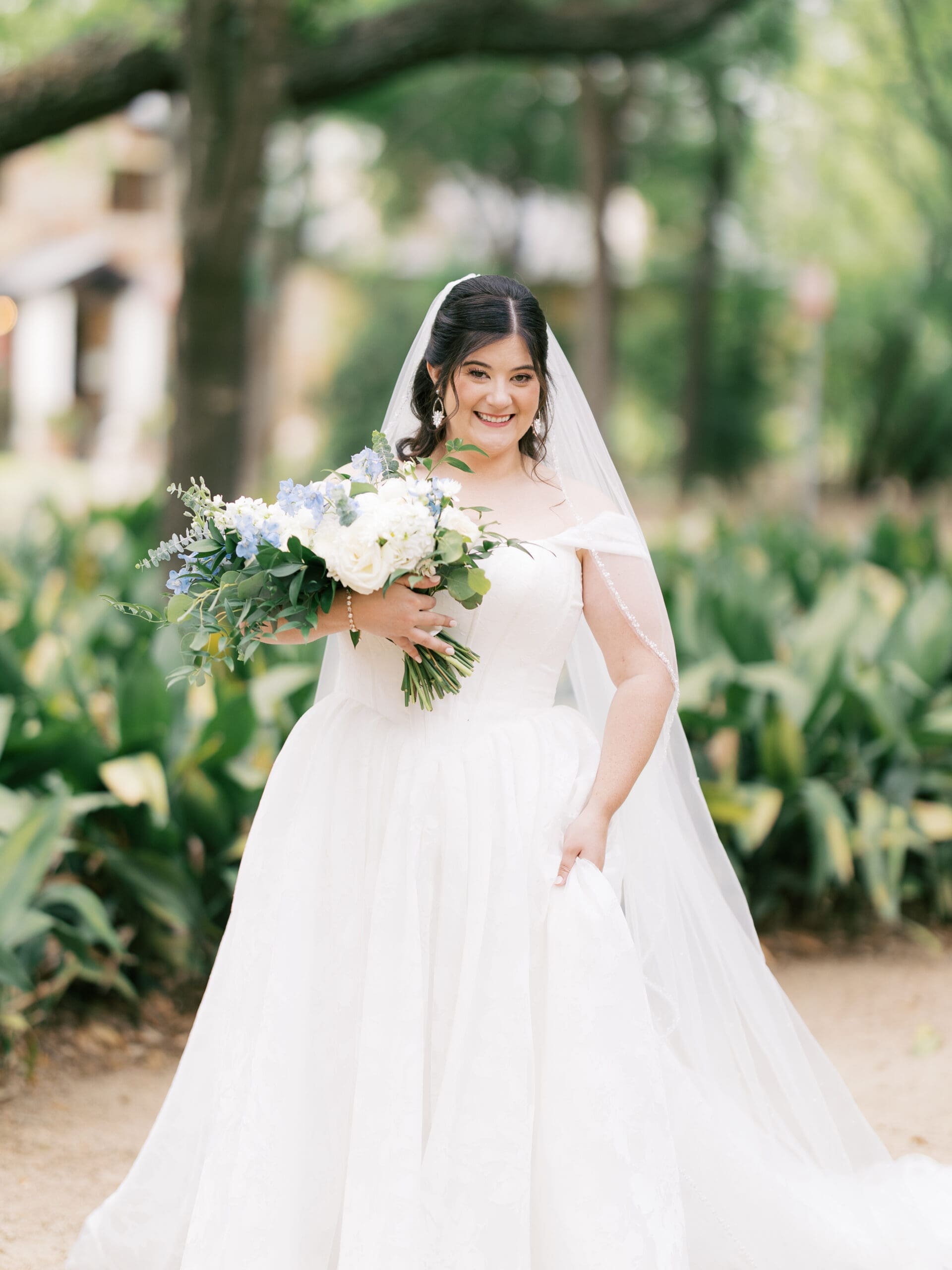 Bride smiling warmly while holding her white and blue bouquet in the lush garden courtyard of the wedding venue.
