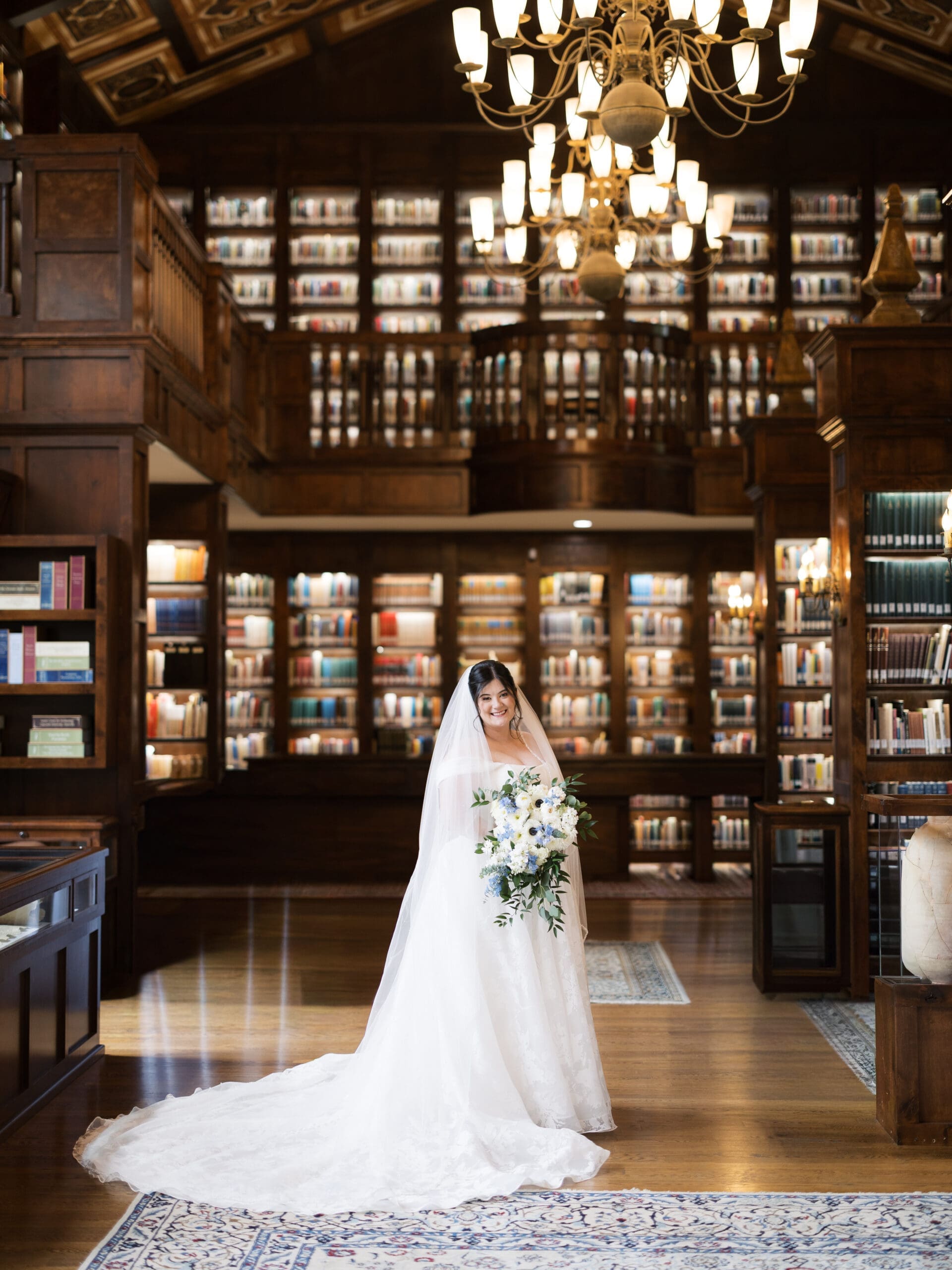 Full-length bridal portrait of a bride standing inside the grand two-story library hall at The Lanier Theological Library, her cathedral veil and lace gown pooling around her beneath a glowing chandelier.
