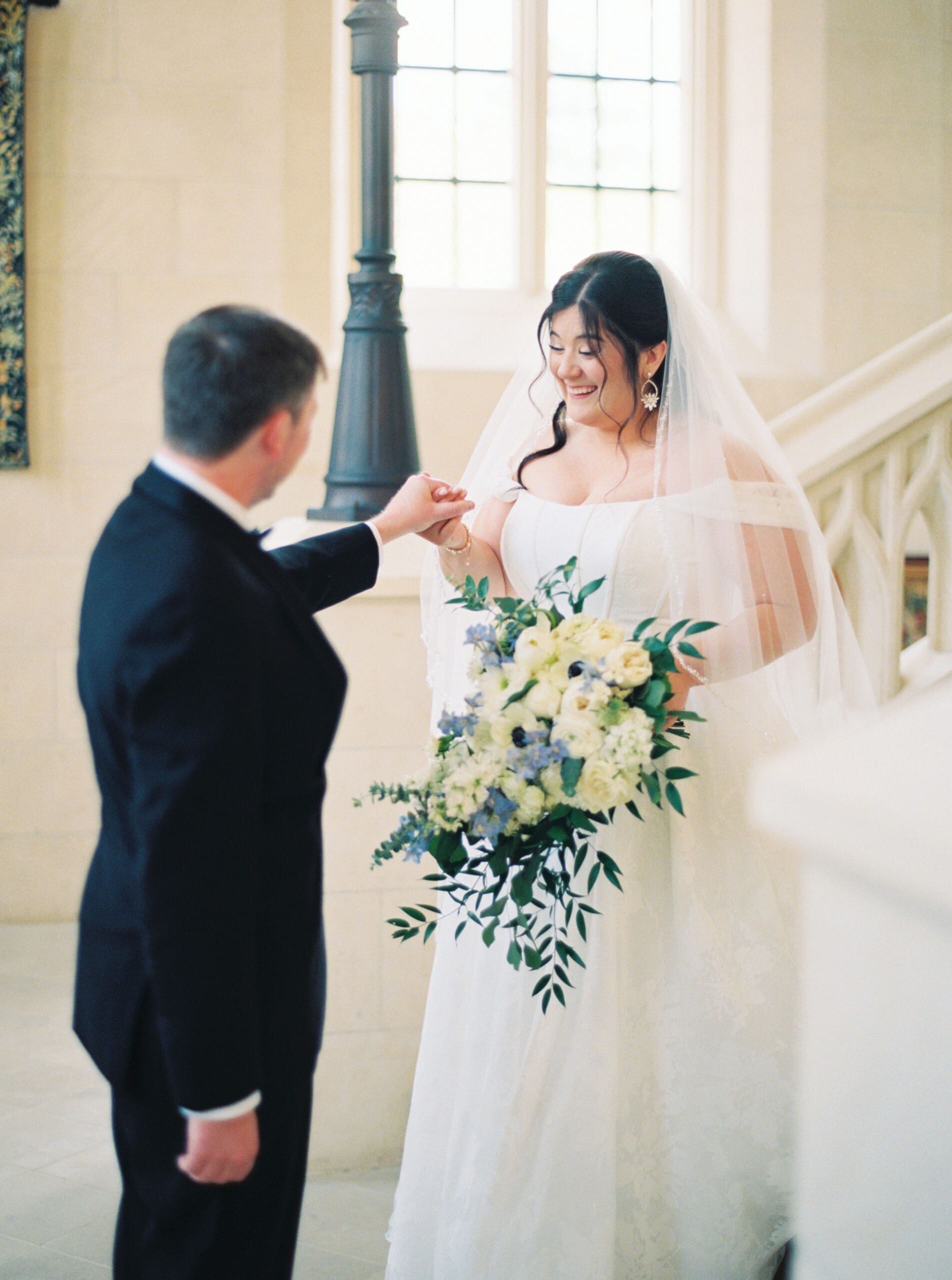 Bride laughing joyfully as the groom holds her hand during their first look on the grand staircase.
