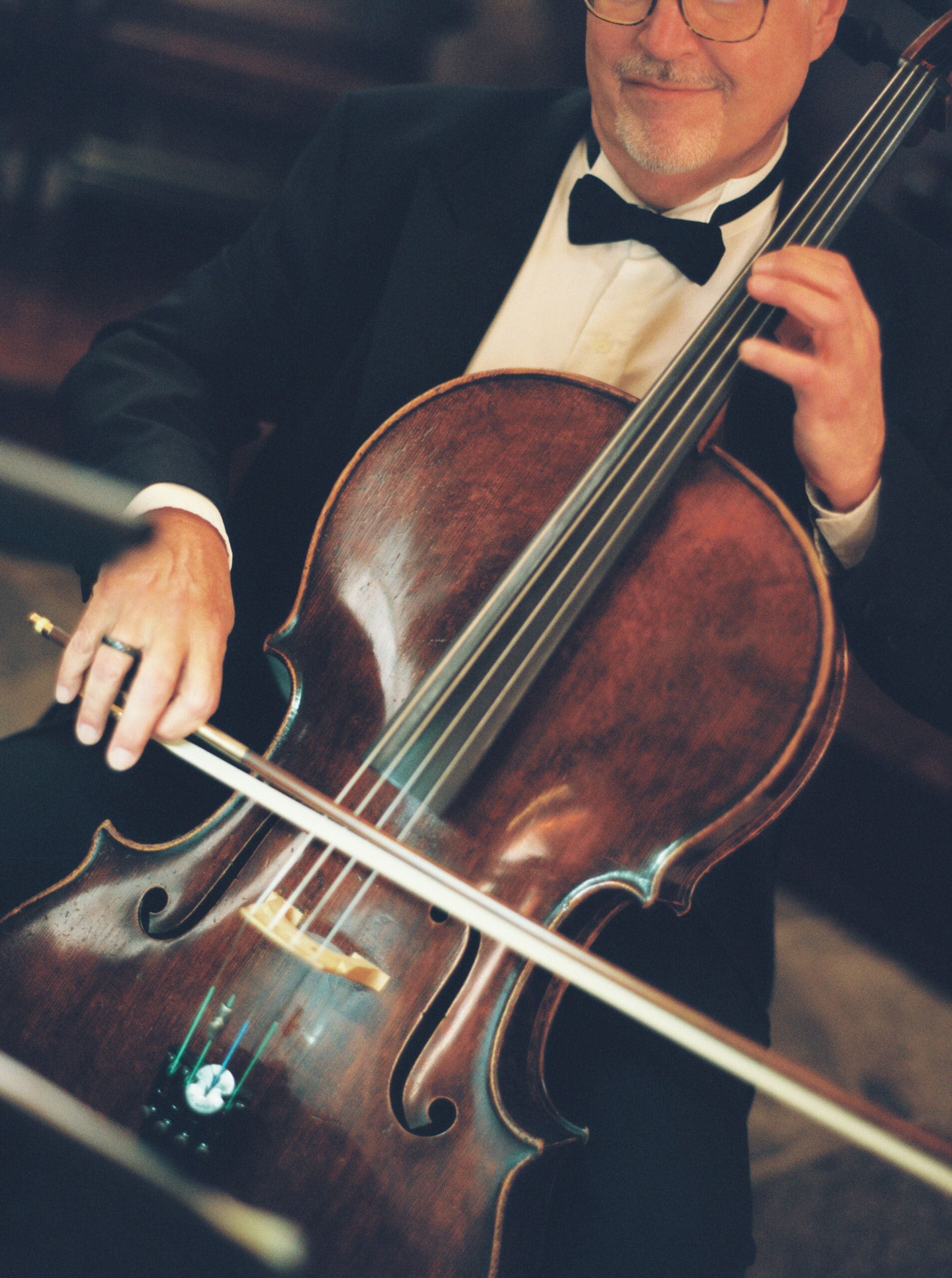 Close-up of a tuxedoed cellist performing during the wedding ceremony, with the warm wood of his instrument filling the frame.