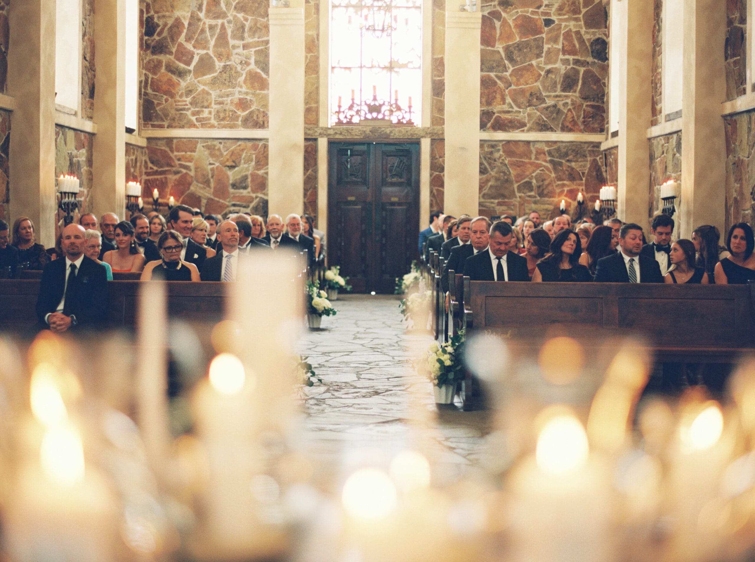 Wedding guests seated in wooden pews inside the stone chapel, photographed through glowing candles in the foreground as they await the ceremony.