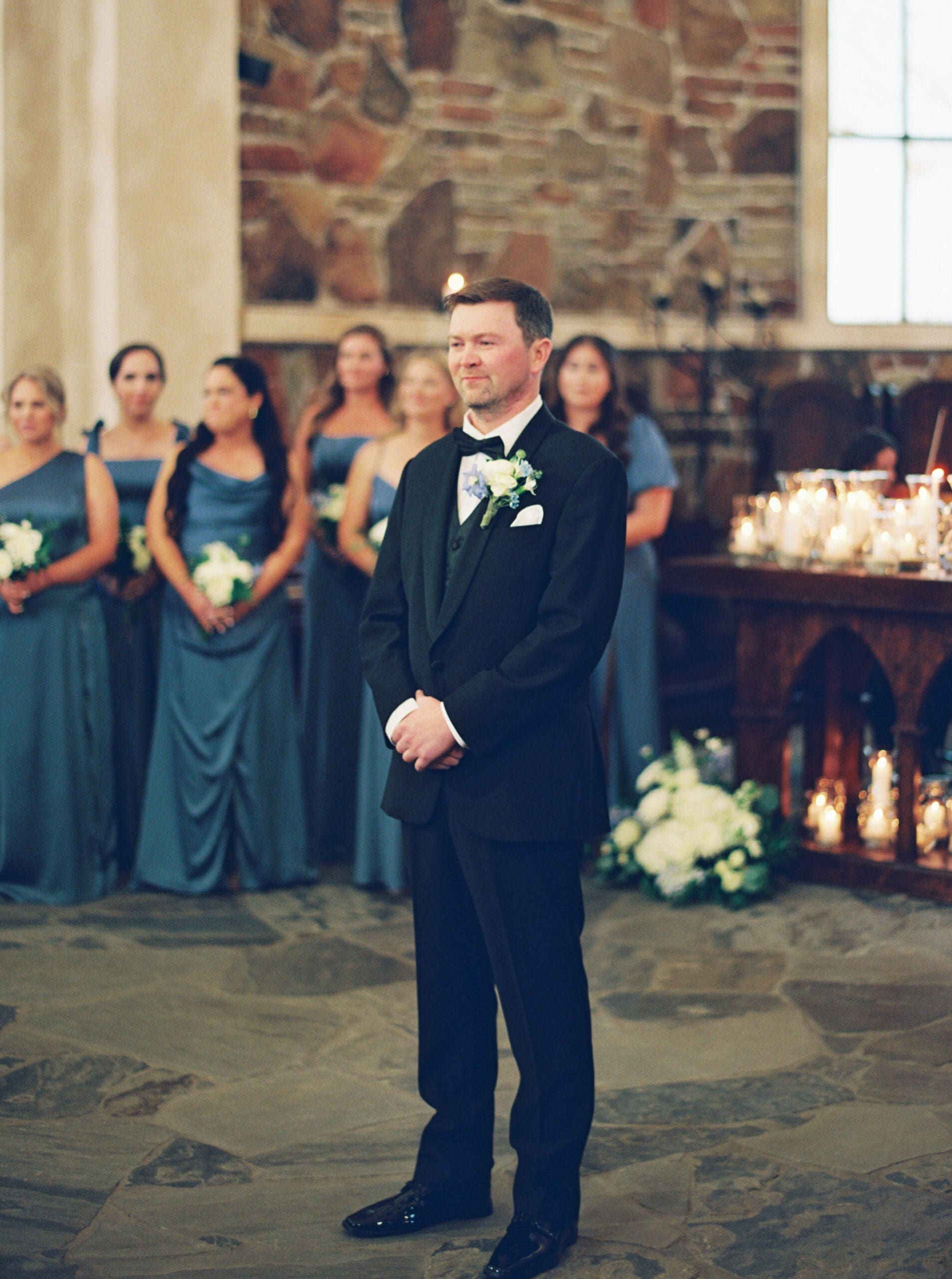 Groom standing at the altar of The Lanier Theological Library chapel with an emotional smile, blue-gowned bridesmaids and candlelit altar visible behind him.