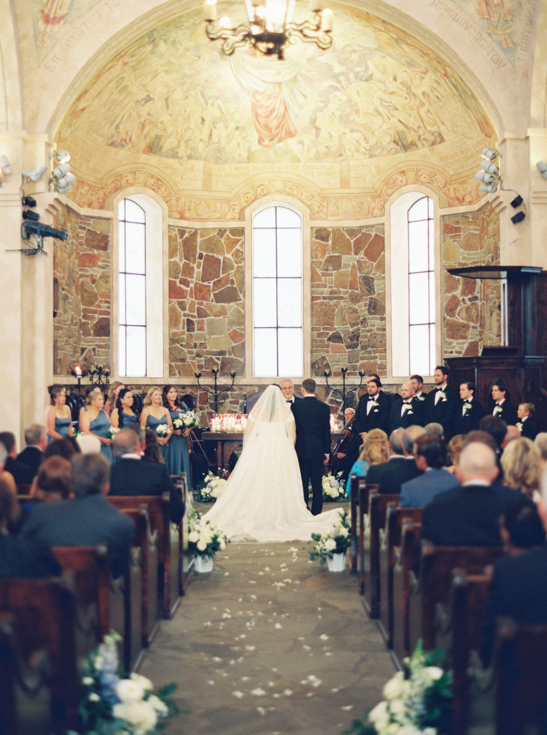 Wide shot of the bride and groom exchanging vows inside the chapel at The Lanier Theological Library, with dusty blue bridesmaids, candlelit altar, and a stunning painted dome above.