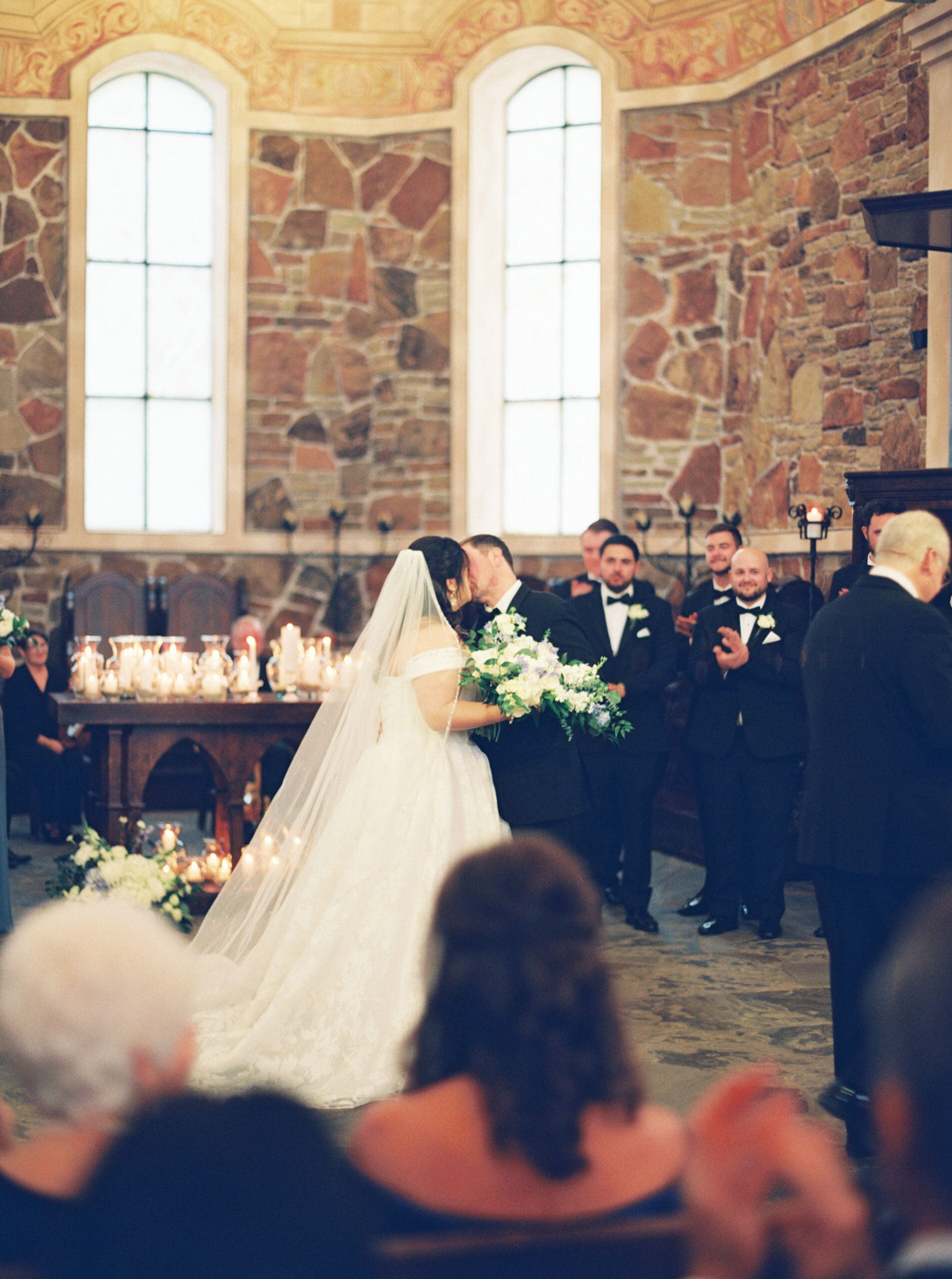 Bride and groom sharing their first kiss at the altar inside the stone chapel, surrounded by candlelight and cheering groomsmen.
