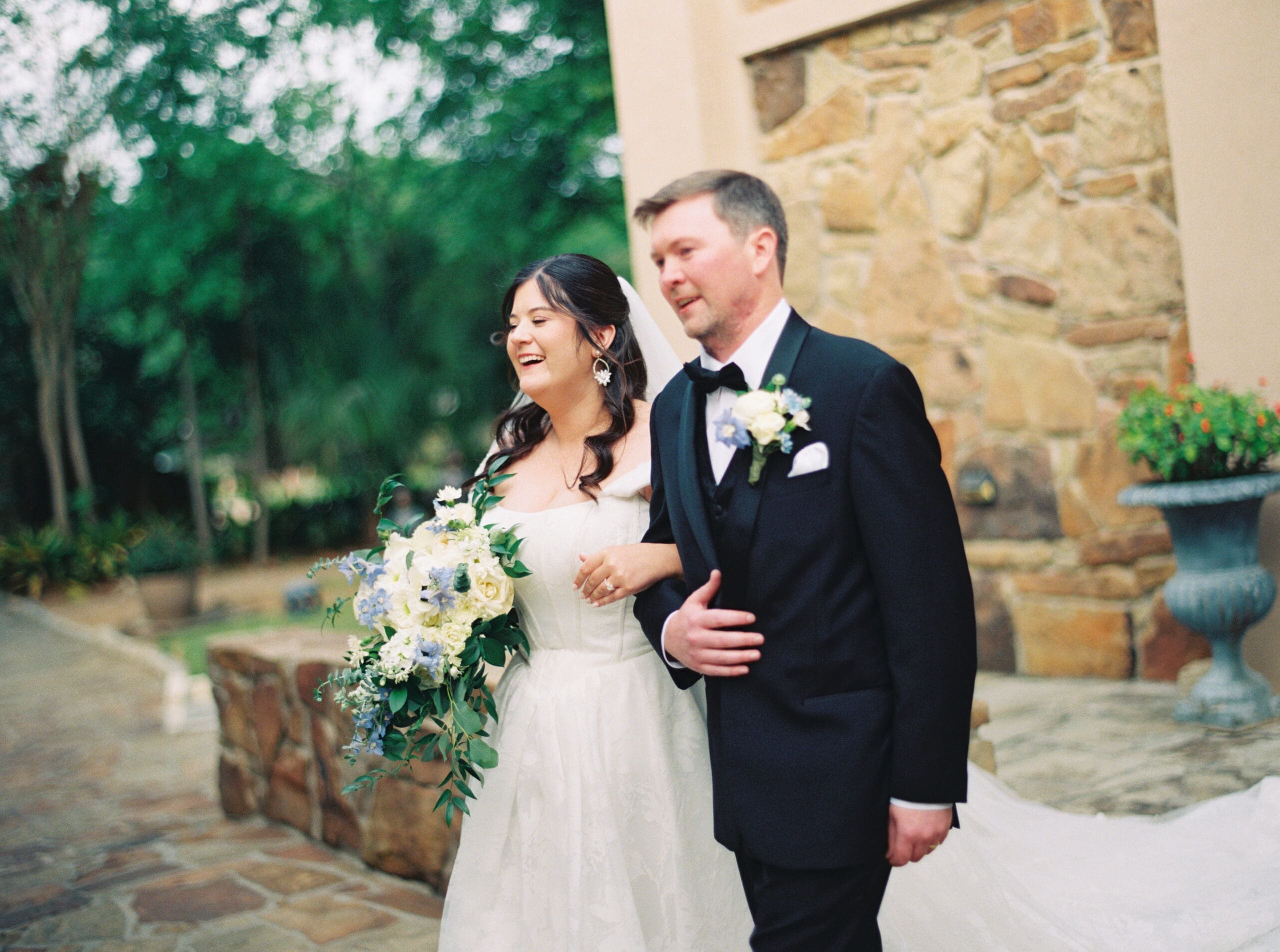 Bride laughing joyfully while walking arm in arm with her groom along the stone courtyard of their wedding venue.
