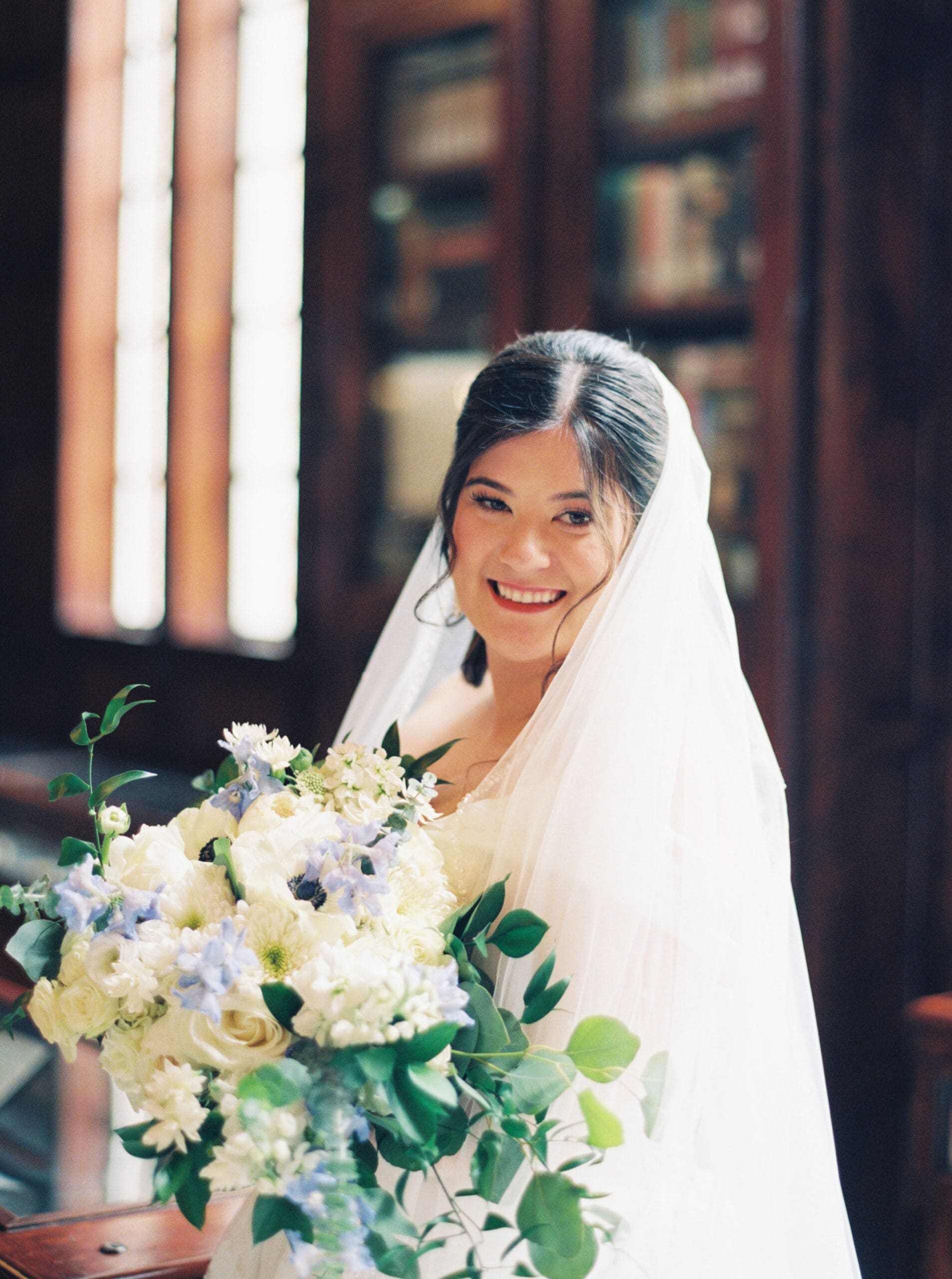 Radiant bride holding a lush white and blue bouquet inside the warm, book-lined interior of the library wedding venue.
