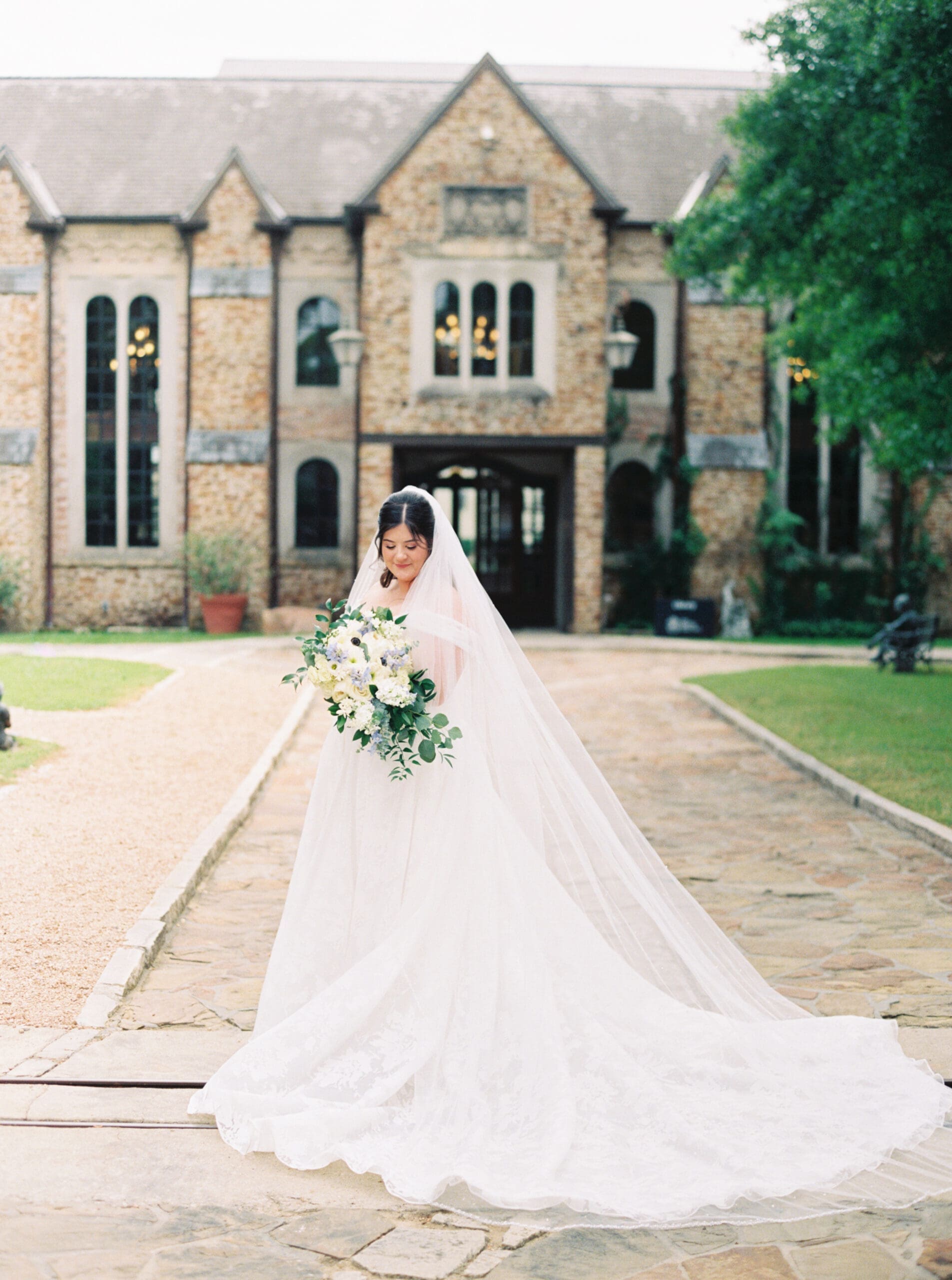 Bride looking down at her bouquet while standing in front of the stone entrance of The Lanier Theological Library, her cathedral veil and lace train sweeping across the cobblestone courtyard.
