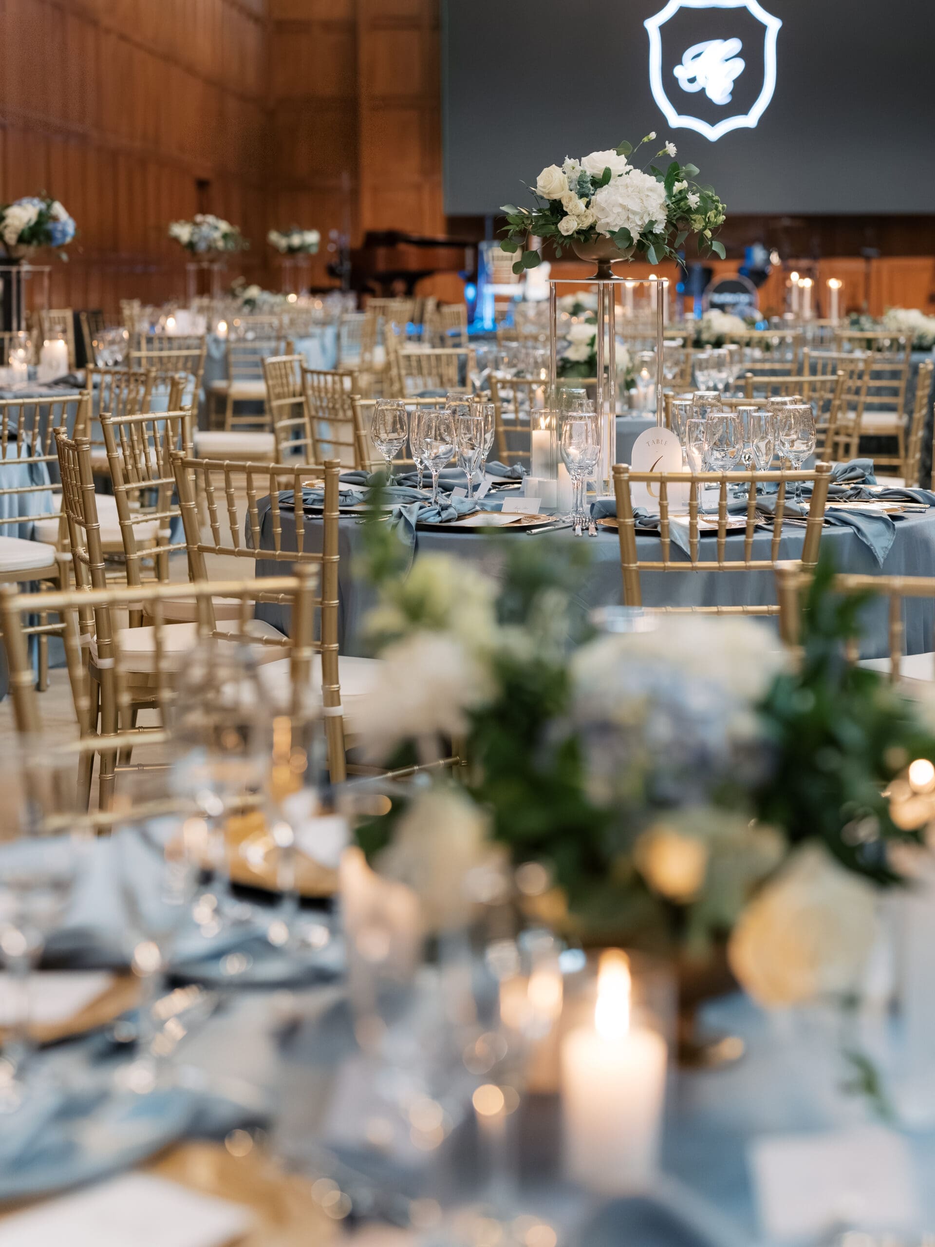 Wide shot of the elegantly styled wedding reception hall at The Lanier Theological Library, featuring dusty blue linens, gold chiavari chairs, candlelight, and white floral centerpieces.
