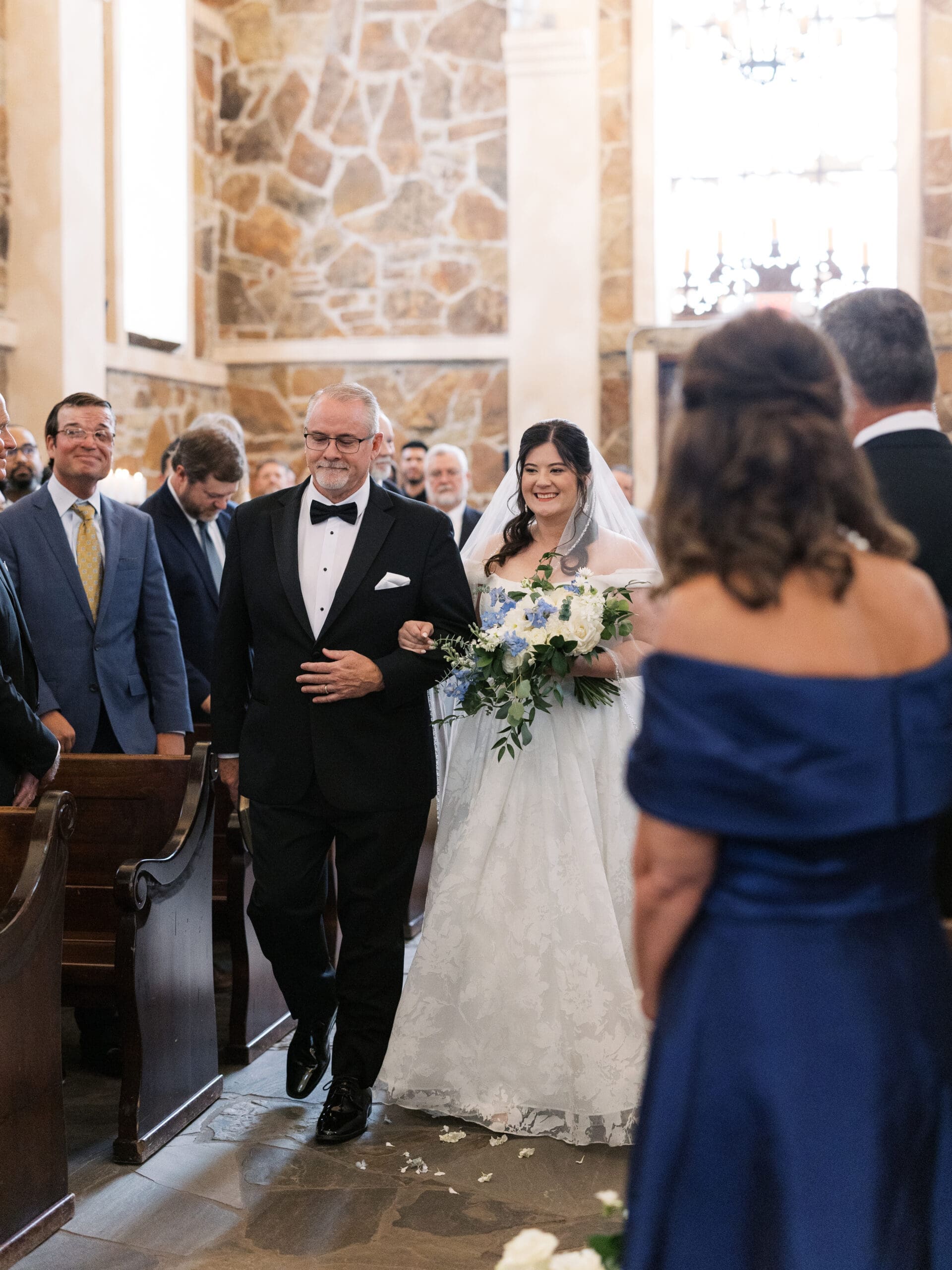 Father of the bride proudly walking his daughter down the aisle of the stone chapel, with guests looking on from wooden pews.