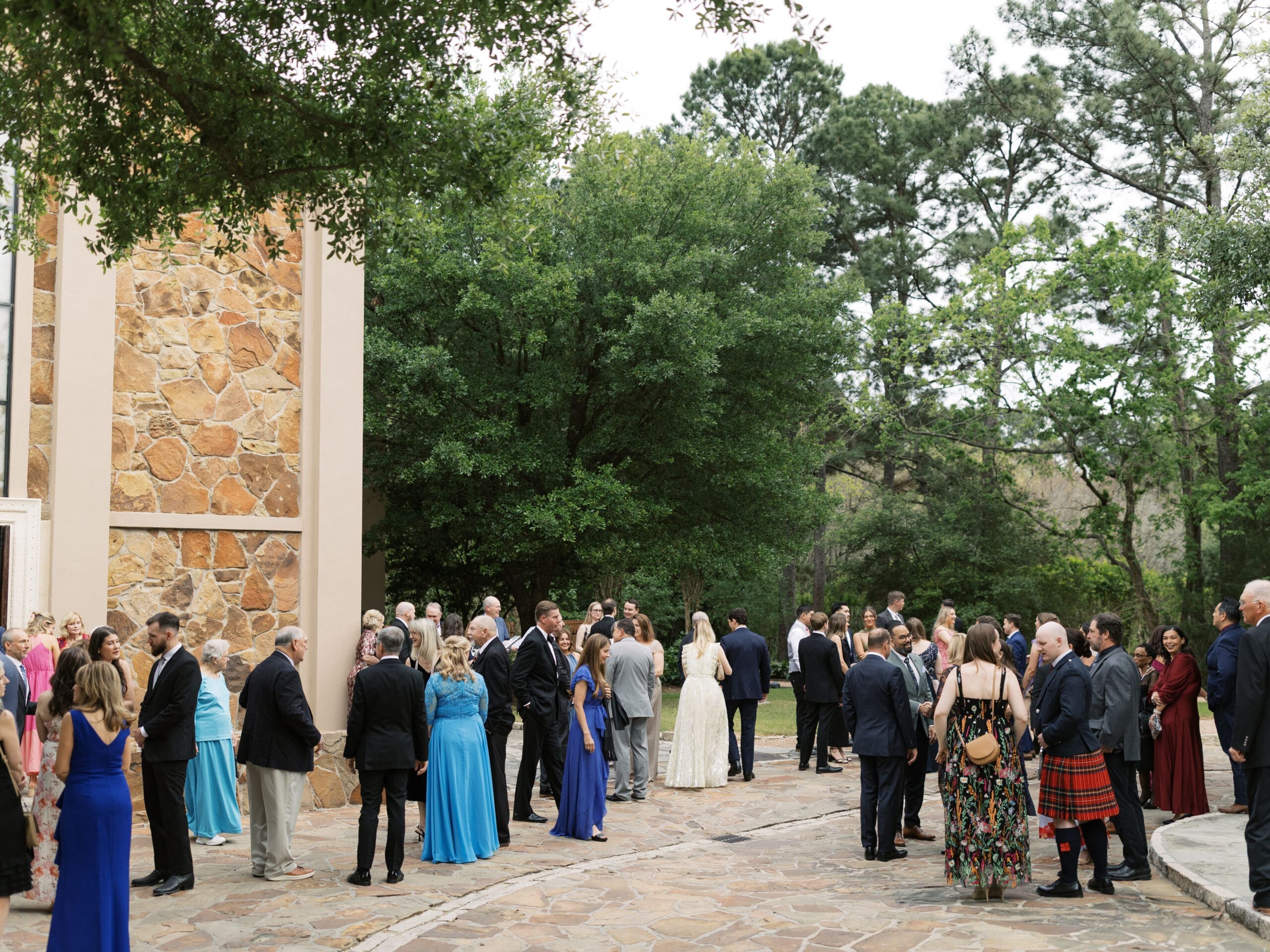Wedding guests mingling on the stone courtyard outside The Lanier Theological Library after the ceremony, surrounded by towering oak and pine trees.
