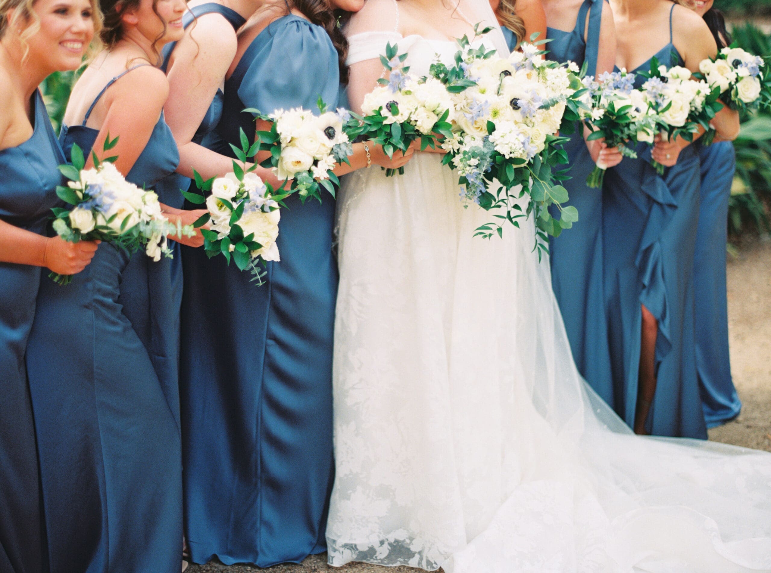 Close-up of the bridal party huddled together holding white and blue bouquets, their dusty blue satin gowns contrasting beautifully against the bride's lace gown at The Lanier Theological Library.
