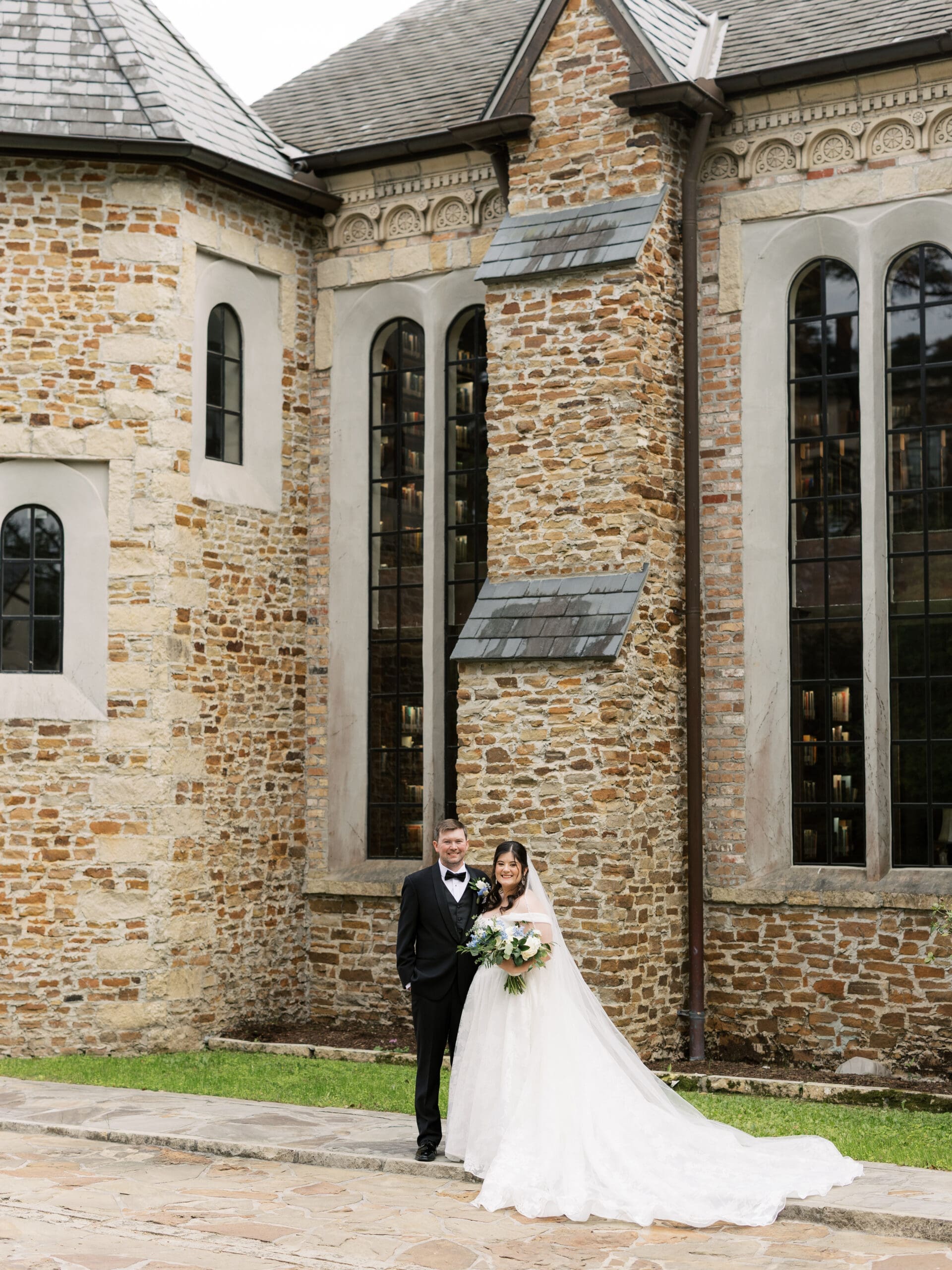 Bride and groom smiling together for a portrait in front of the ivy-covered stone facade of The Lanier Theological Library, her lace train cascading across the flagstone.
