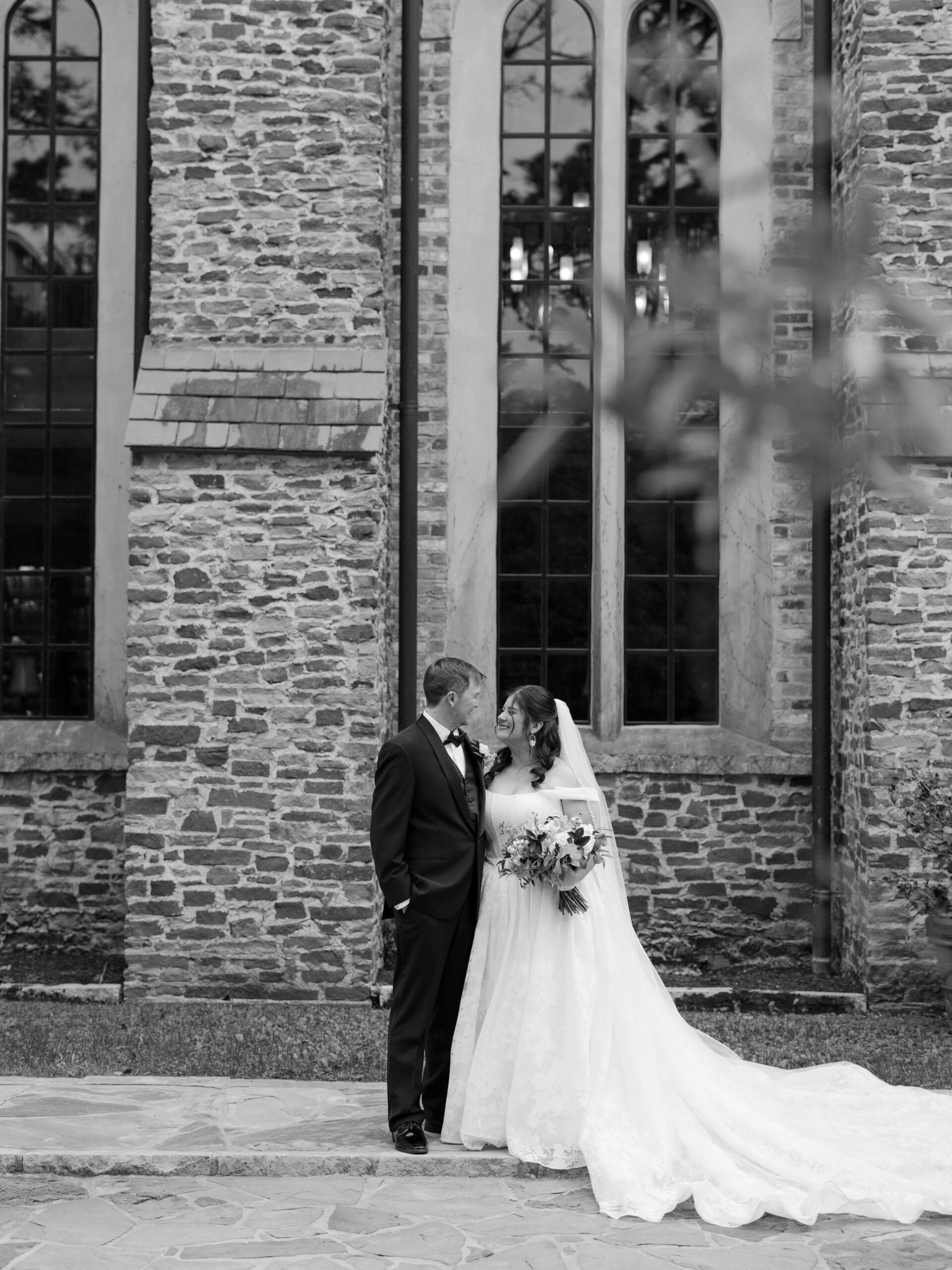 Black and white portrait of a bride laughing with her groom against the stone exterior wall of the wedding venue.
