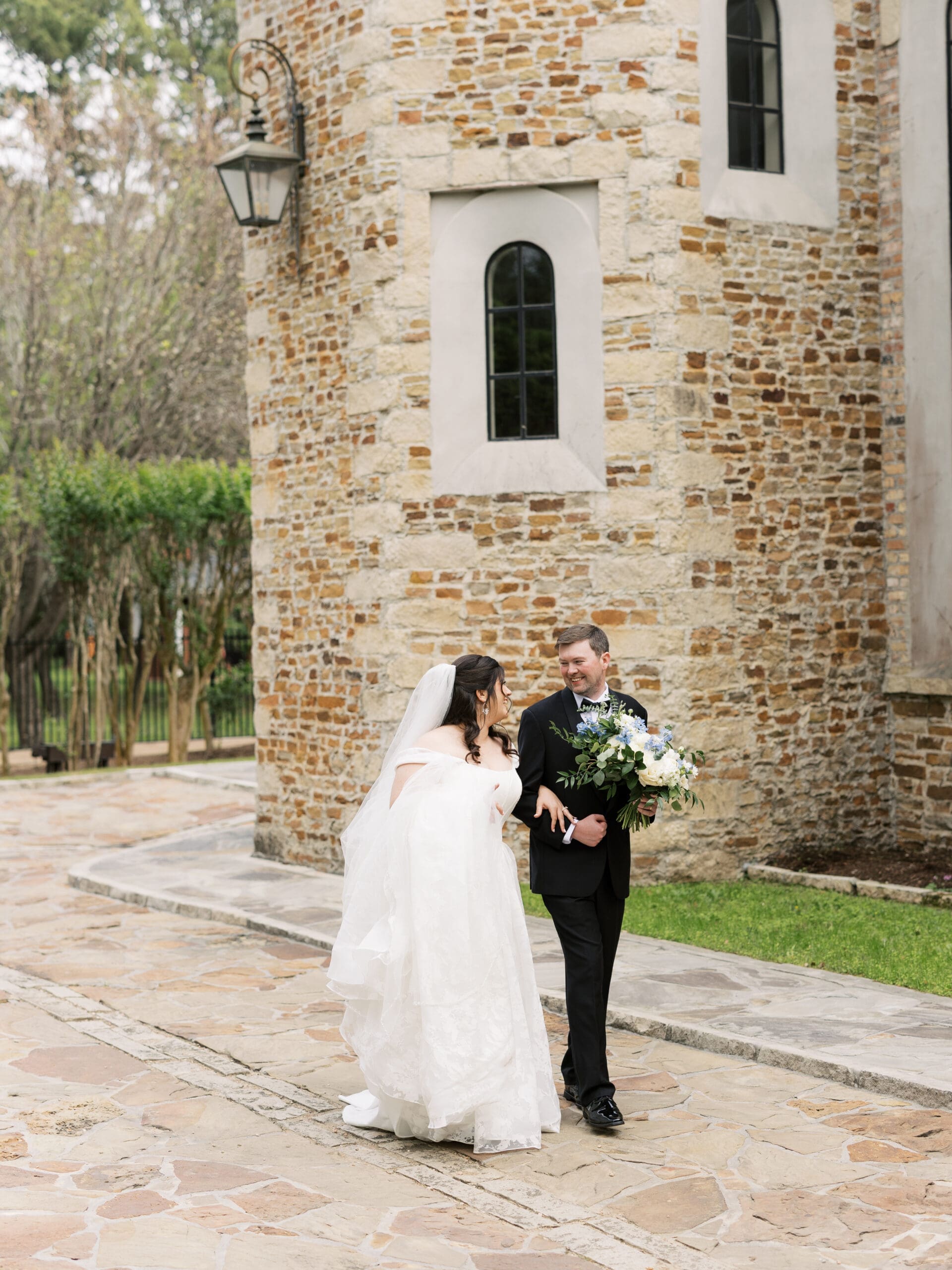Bride and groom laughing together as they walk along the stone courtyard beside the rounded chapel tower of The Lanier Theological Library.
