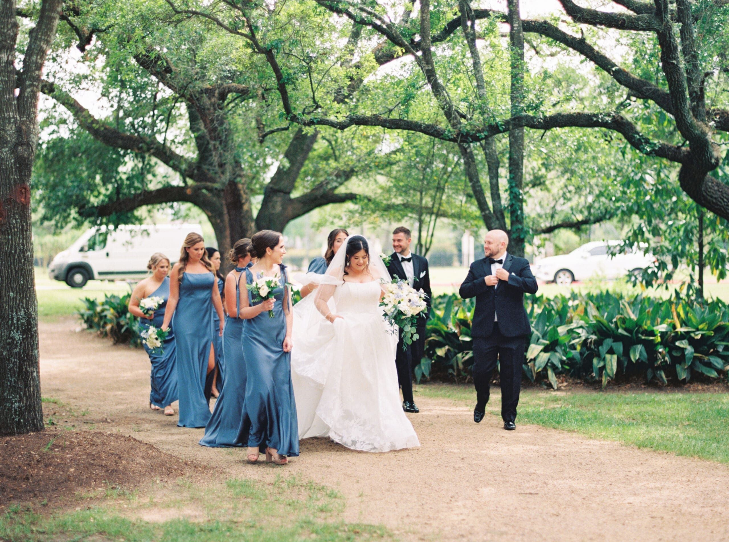 Wedding party walking together beneath a canopy of oak trees on the grounds of the venue, the bride centered among her blue-gowned bridesmaids and tuxedoed groomsmen.
