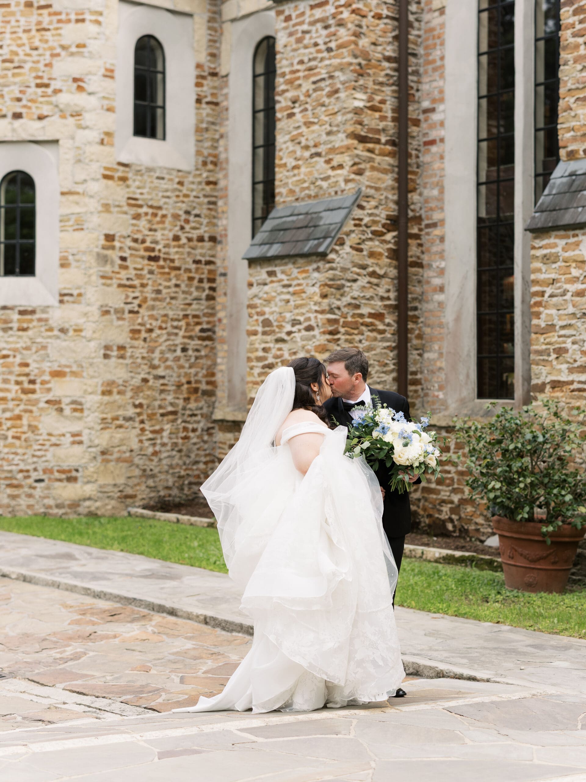 Bride and groom sharing a romantic kiss on the stone courtyard, the bride's cathedral veil billowing behind her against the rustic brick chapel exterior.
