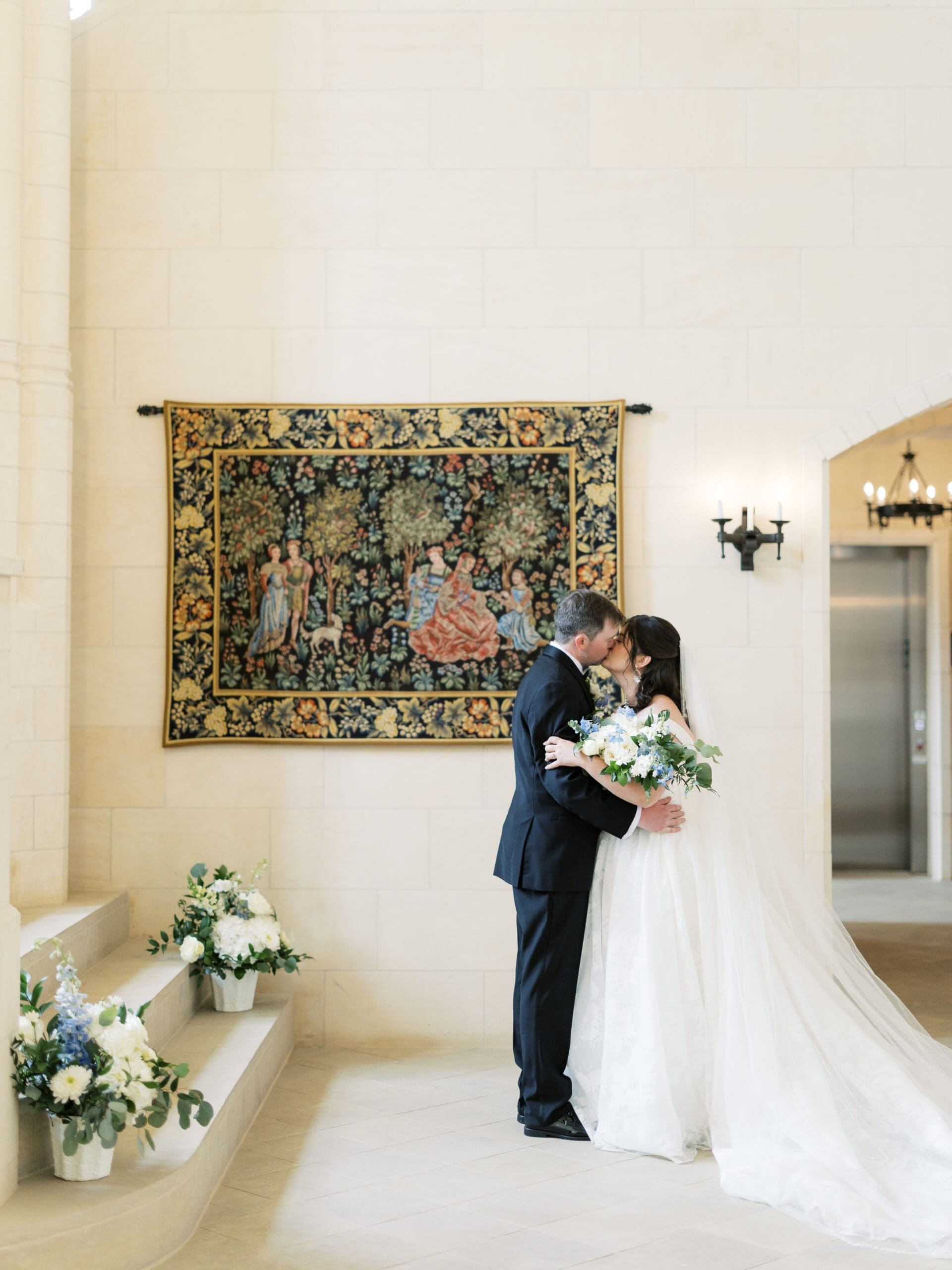 Bride and groom sharing a kiss in the grand entry hall of The Lanier Theological Library, with white and blue floral arrangements lining the steps beside them.
