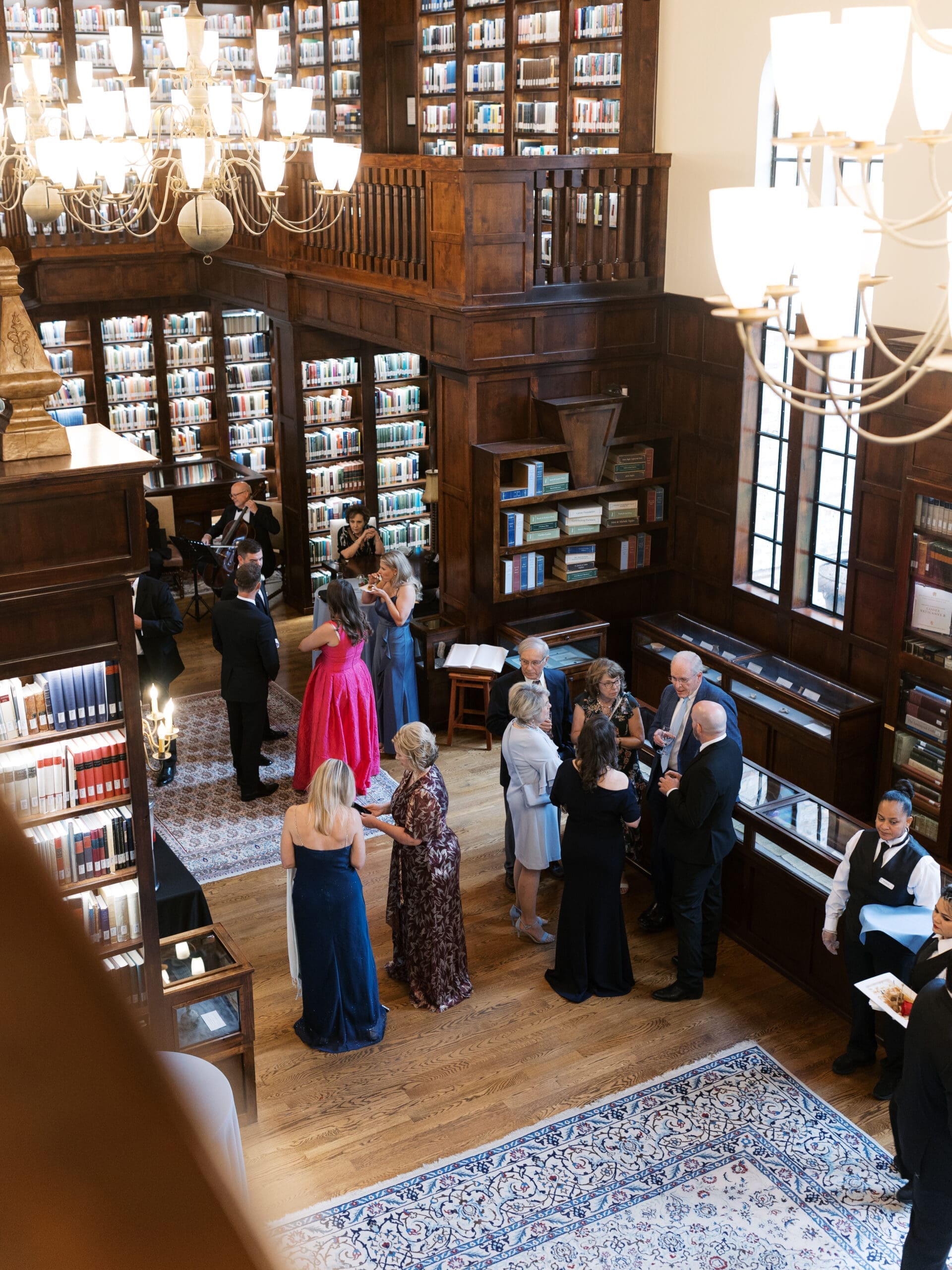 Overhead view of wedding guests mingling in the grand two-story library hall, surrounded by glowing chandeliers and floor-to-ceiling bookshelves.
