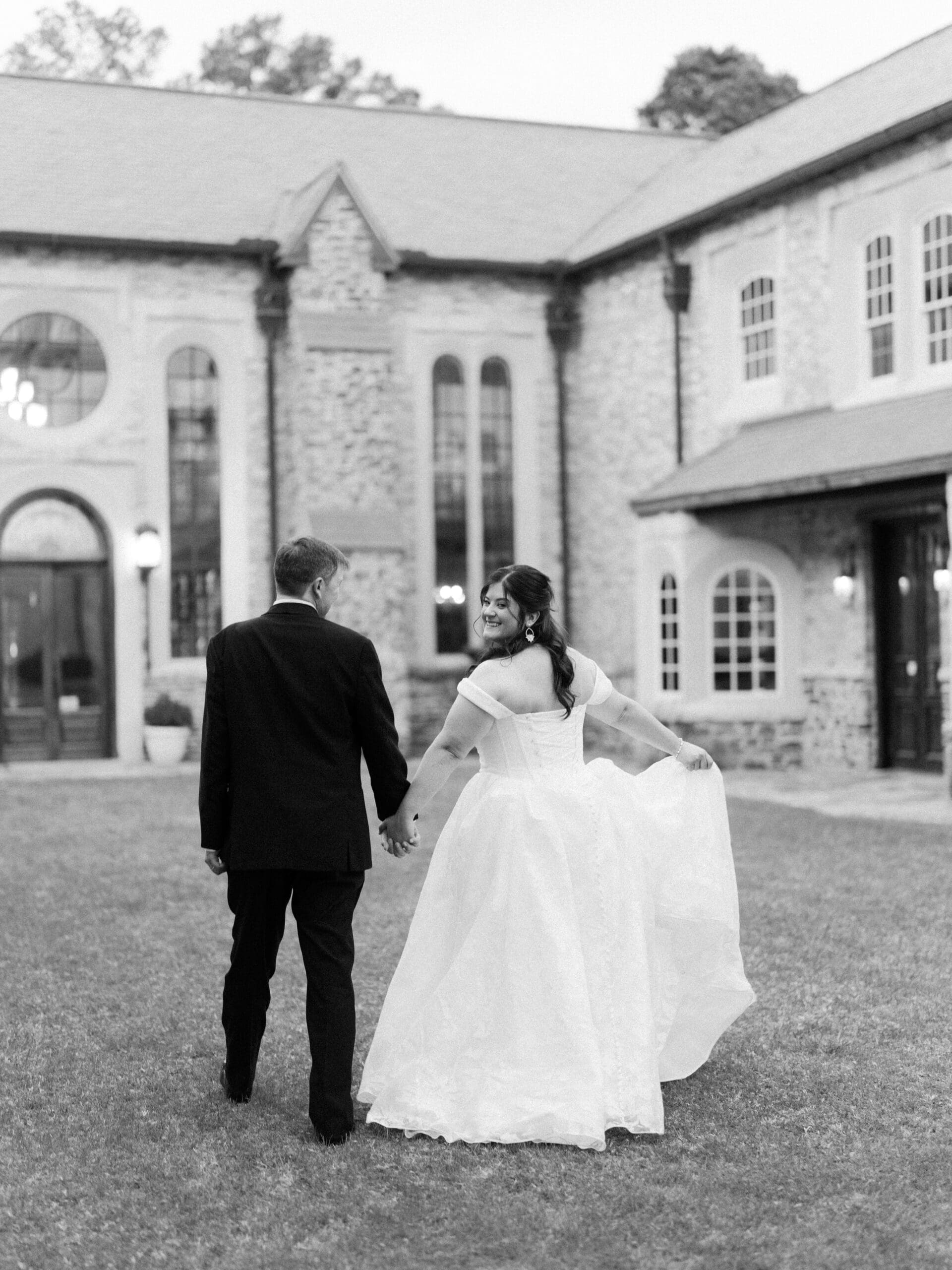 Black and white image of a bride glancing back with a smile as she and her groom walk hand in hand across the lawn at The Lanier Theological Library at dusk.
