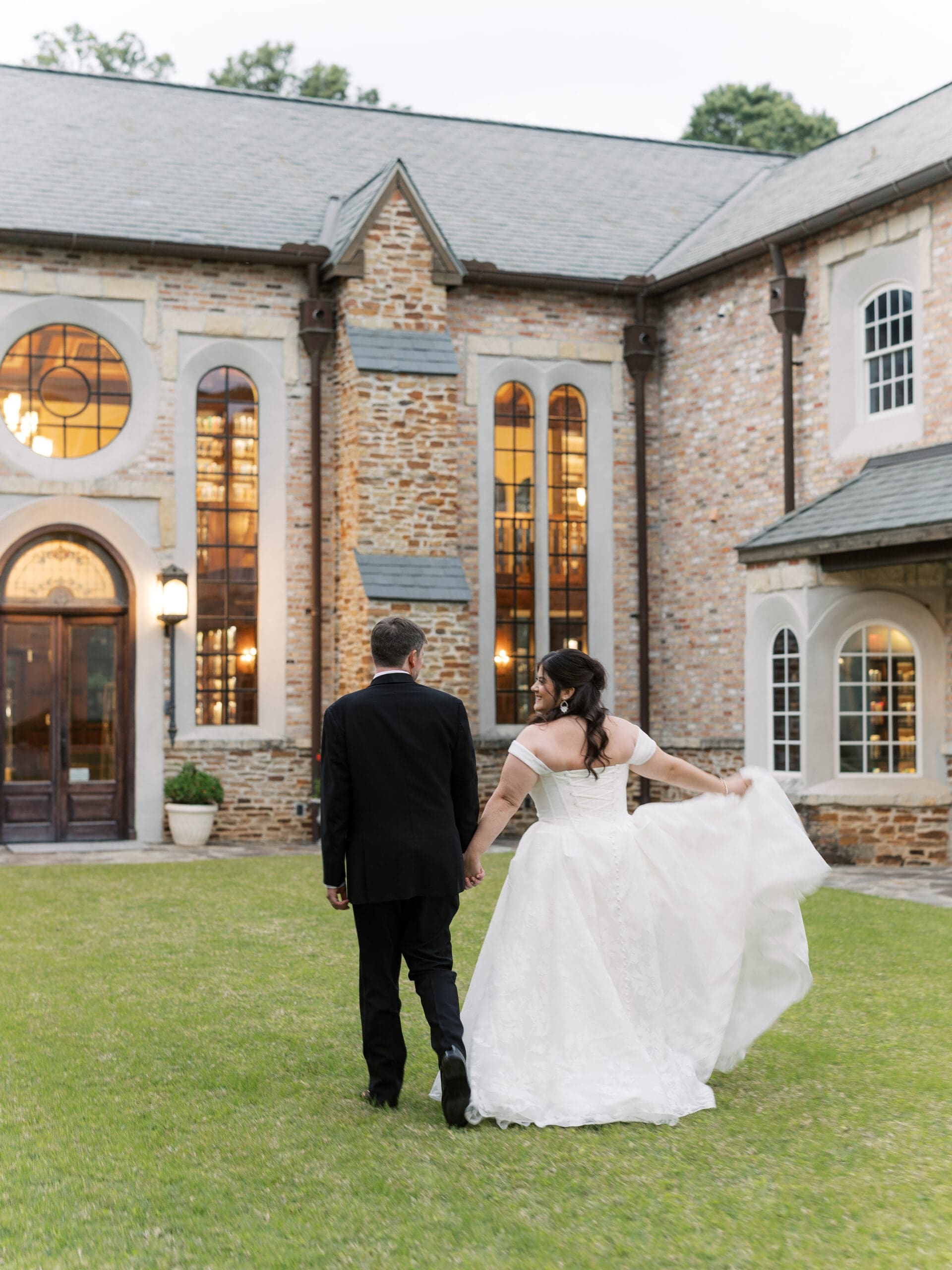 Bride twirling her gown as she and the groom walk hand in hand across the lawn toward the glowing stone building at dusk.
