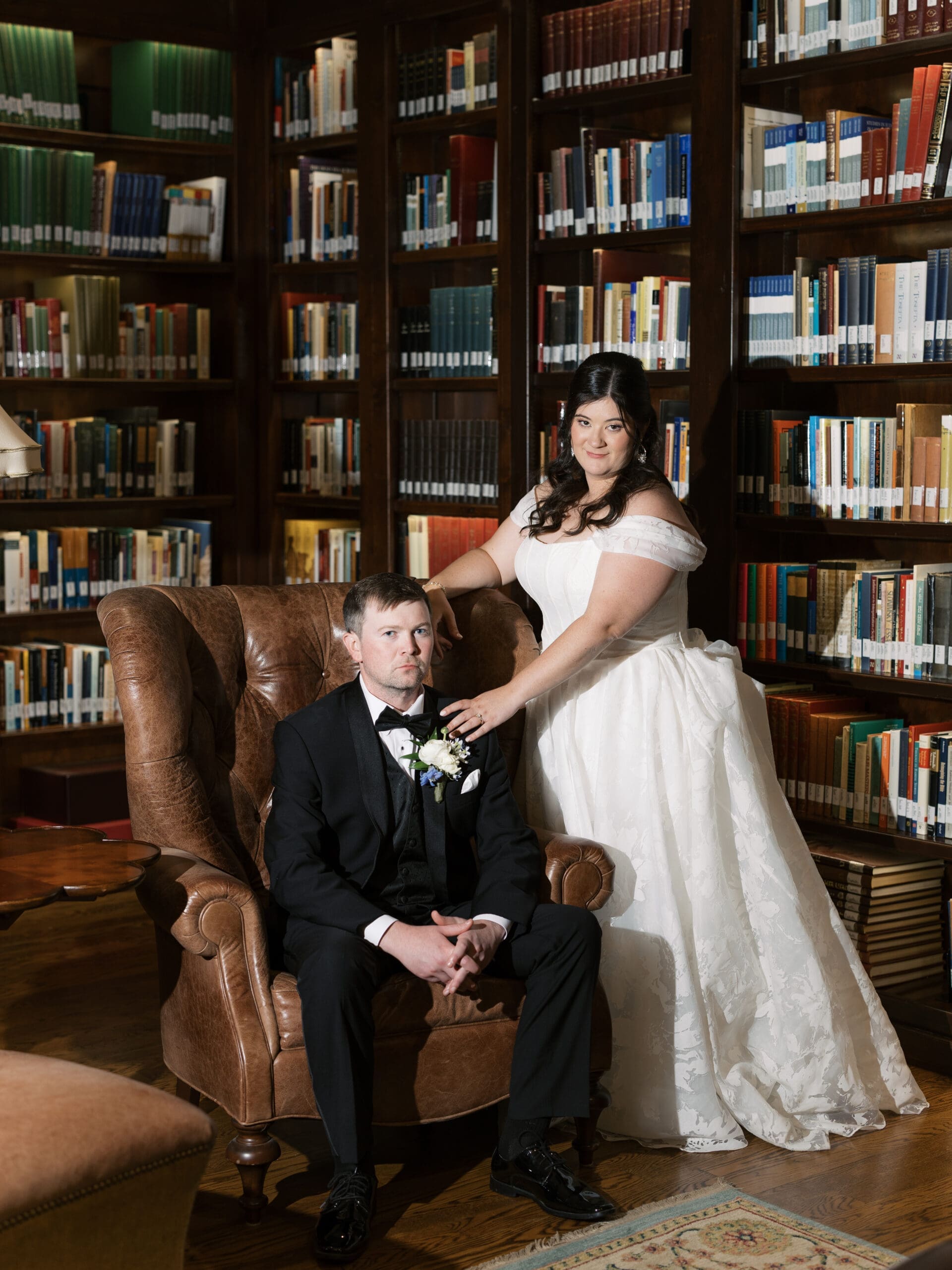 Bride resting her hand on the groom's shoulder as he sits in a leather wingback chair at The Lanier Theological Library, both gazing directly at the camera.
