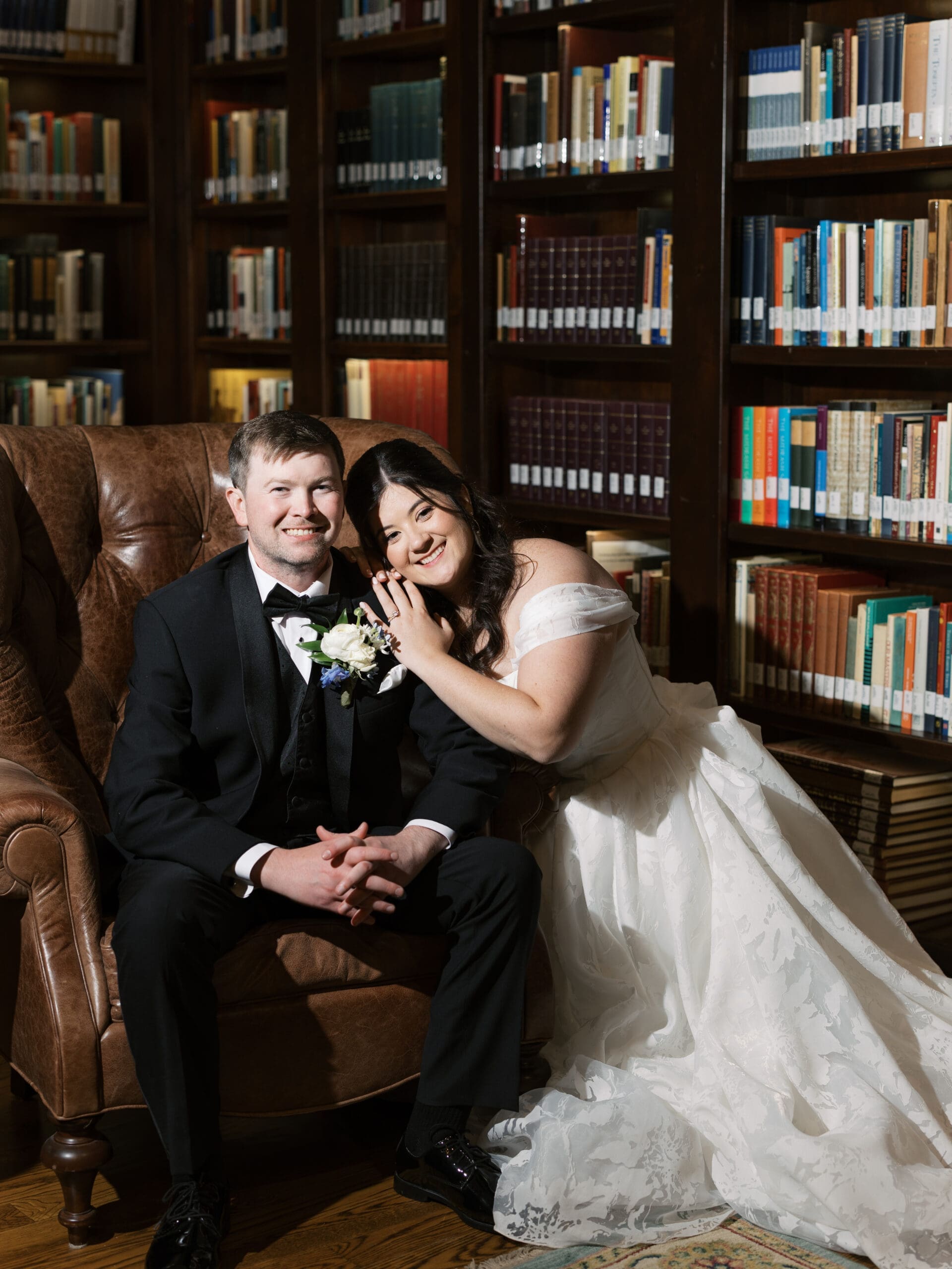 Bride and groom smiling and posing together in a leather armchair at The Lanier Theological Library, surrounded by floor-to-ceiling bookshelves.
