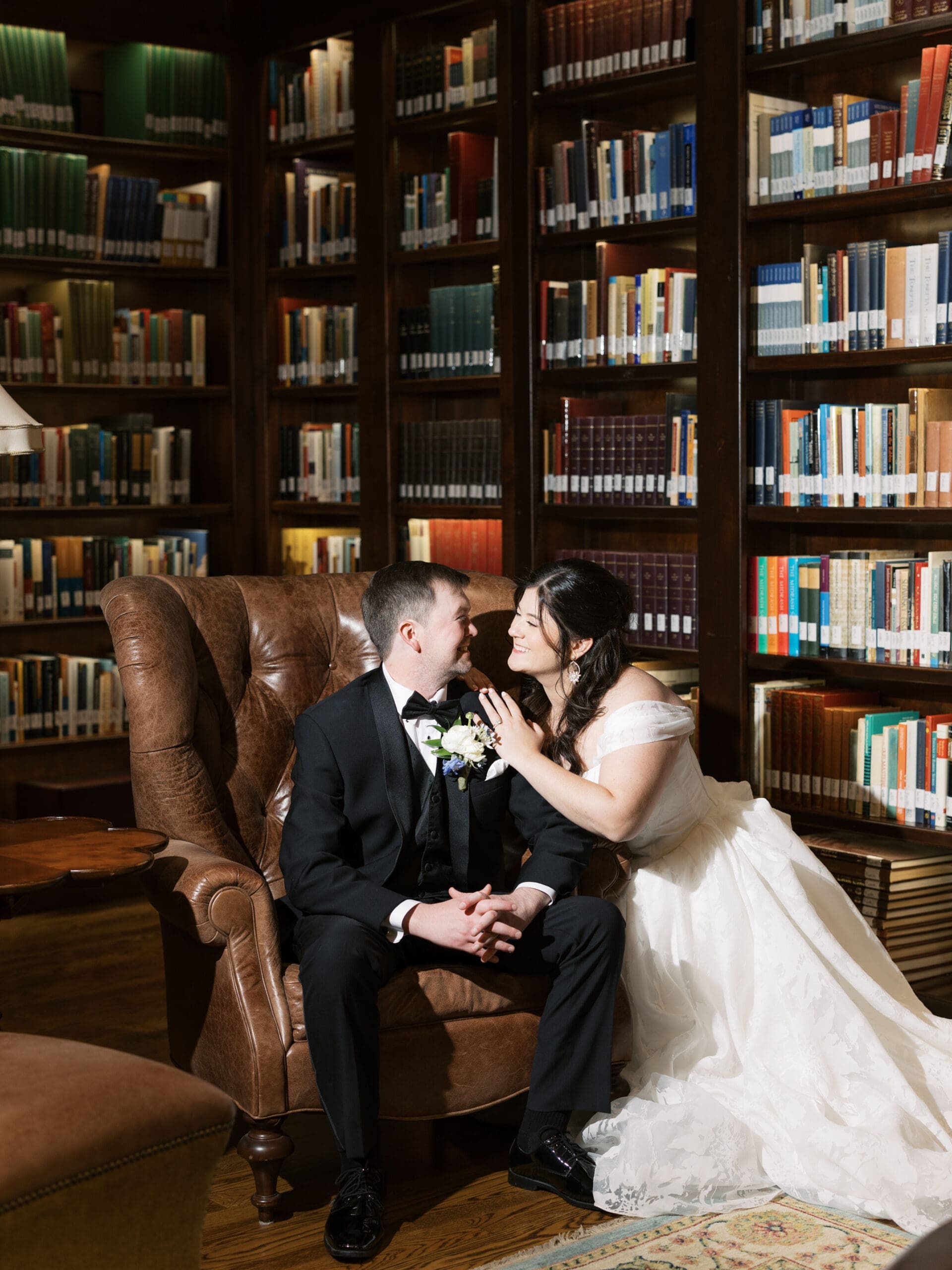 Bride and groom gazing at each other while seated in a leather wingback chair, surrounded by the warm, book-lined walls of the library reception space.