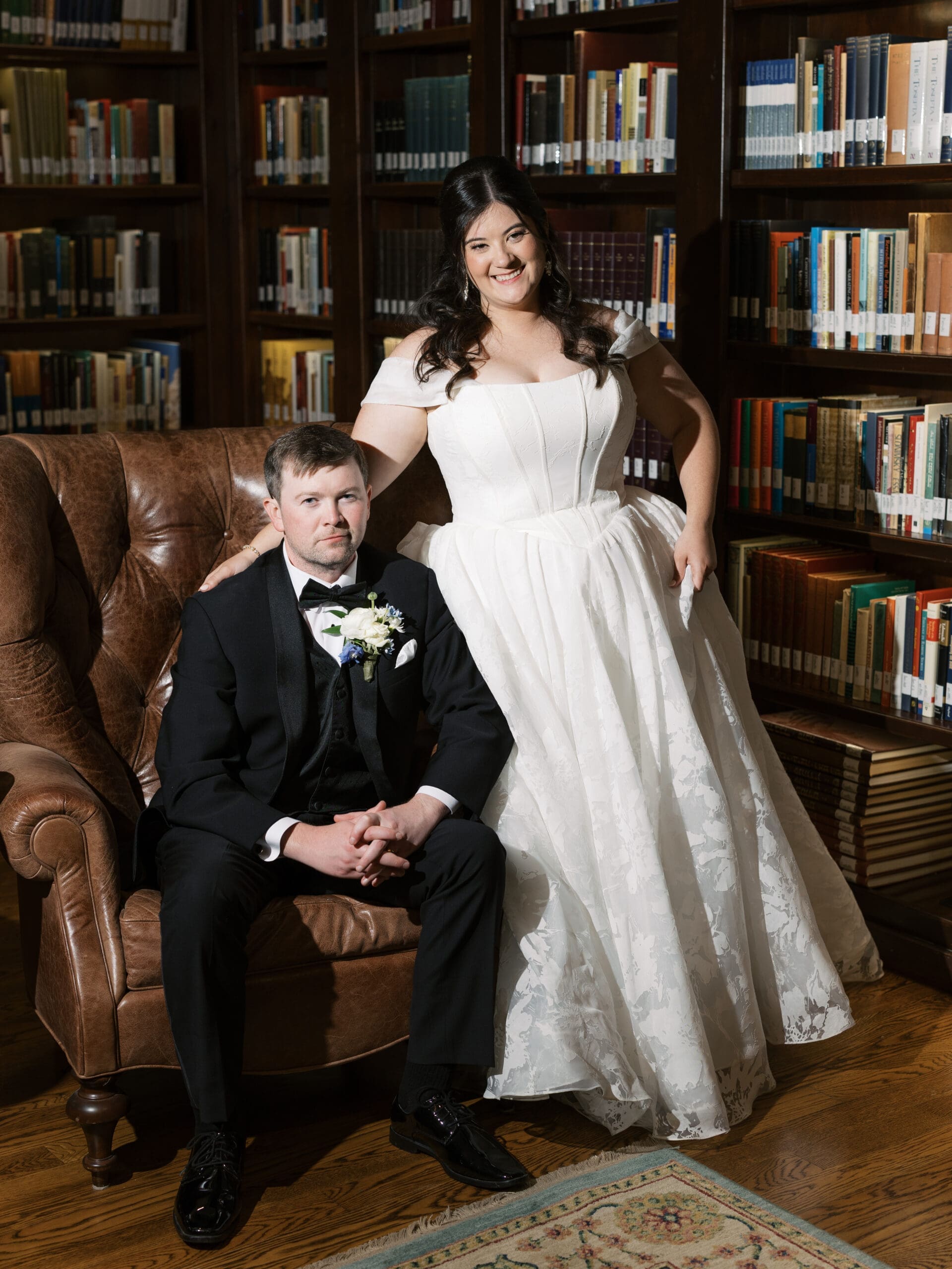 Bride standing confidently beside the groom seated in a leather armchair, both posing in front of the dark wood bookshelves of the library reception space.
