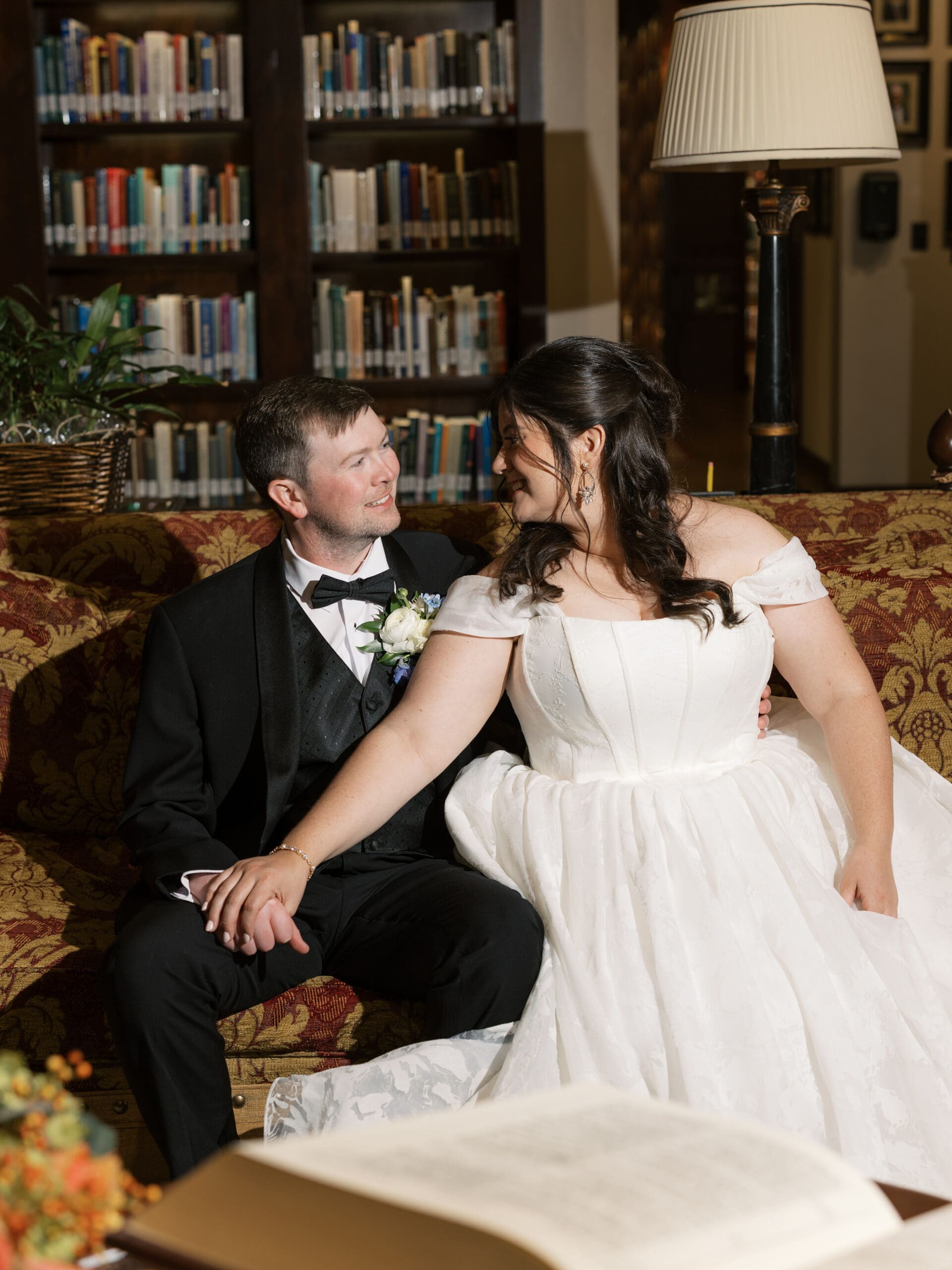 Bride and groom laughing together on a patterned sofa in the cozy reading room of the library reception venue.

