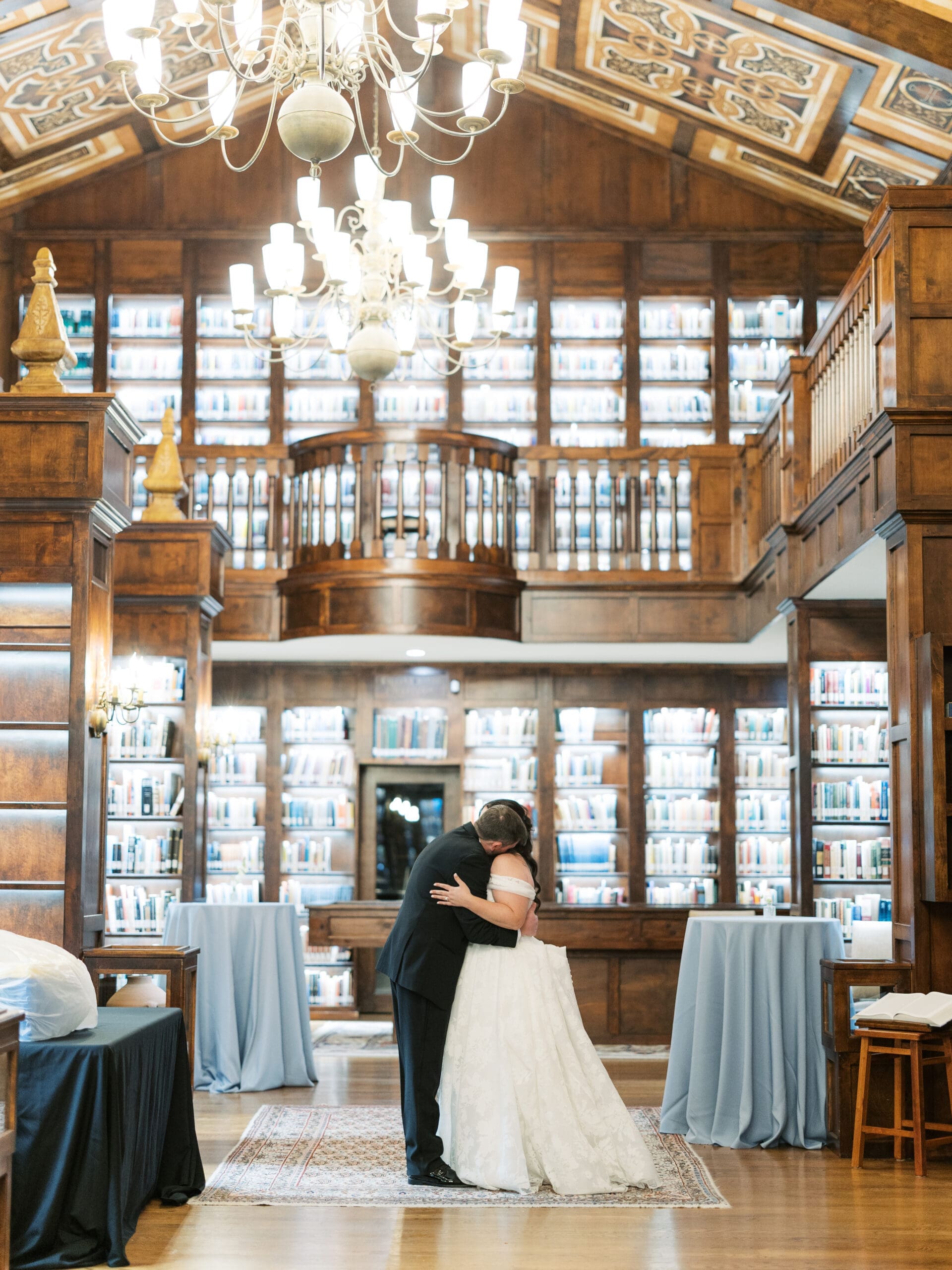 Bride and groom sharing a romantic dip kiss beneath the grand chandelier of The Lanier Theological Library, surrounded by two-story bookshelves and draped reception tables.
