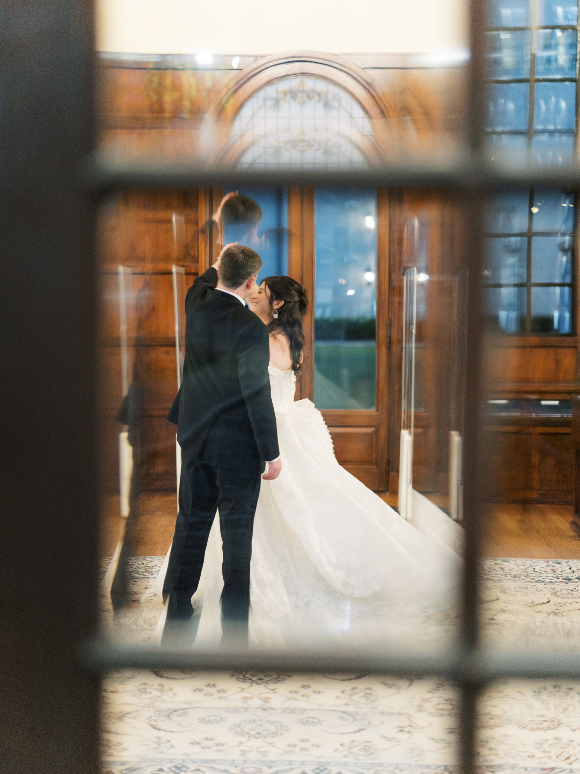 Bride and groom dancing and sharing an intimate moment, captured through the leaded glass windows of the library reception hall.
