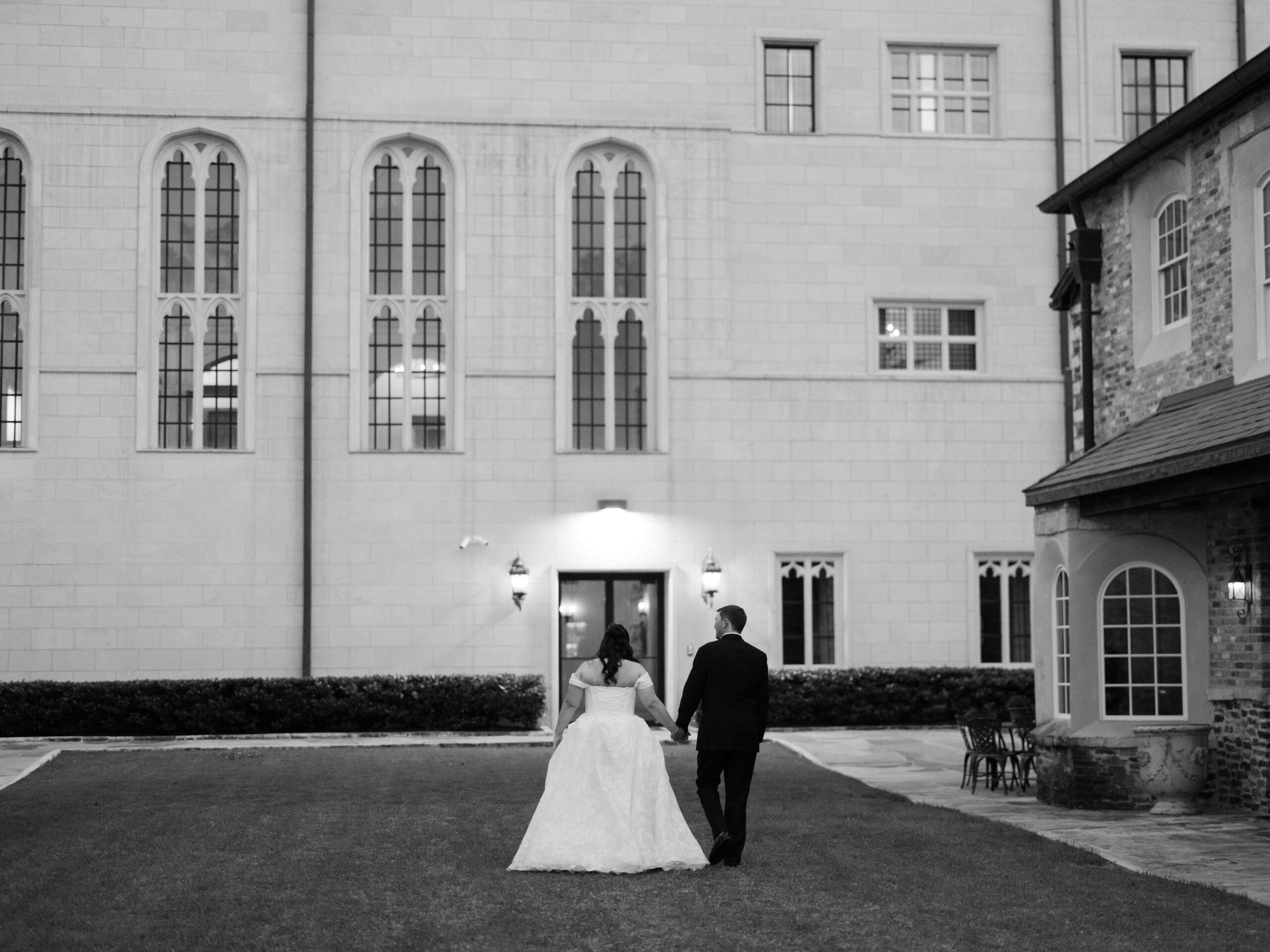 Black and white image of a bride and groom walking hand in hand toward the stone exterior of their wedding venue at night.
