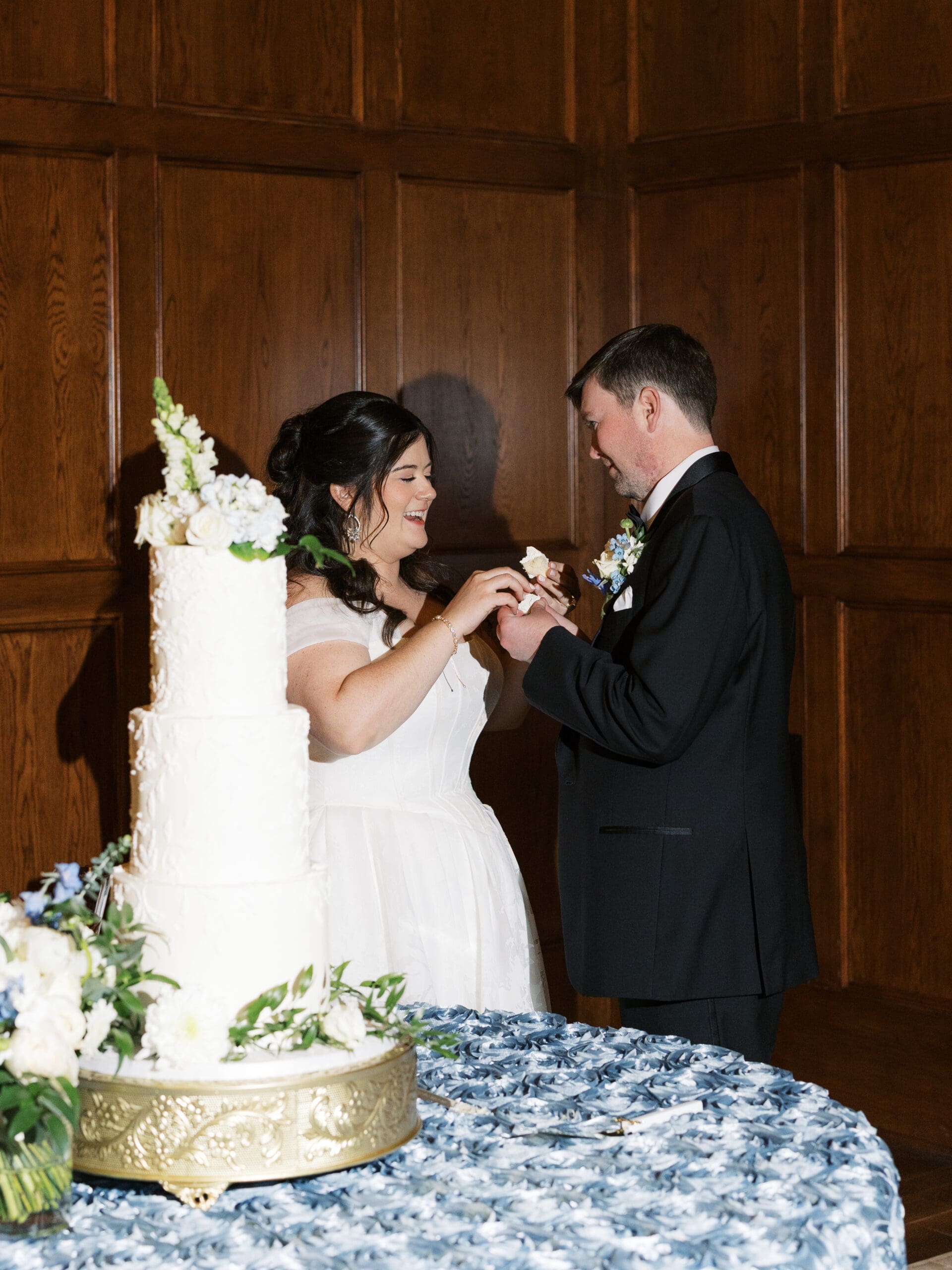 Bride laughing as she feeds the groom a bite of their white tiered wedding cake, decorated with fresh white flowers against a dark wood-paneled wall.
