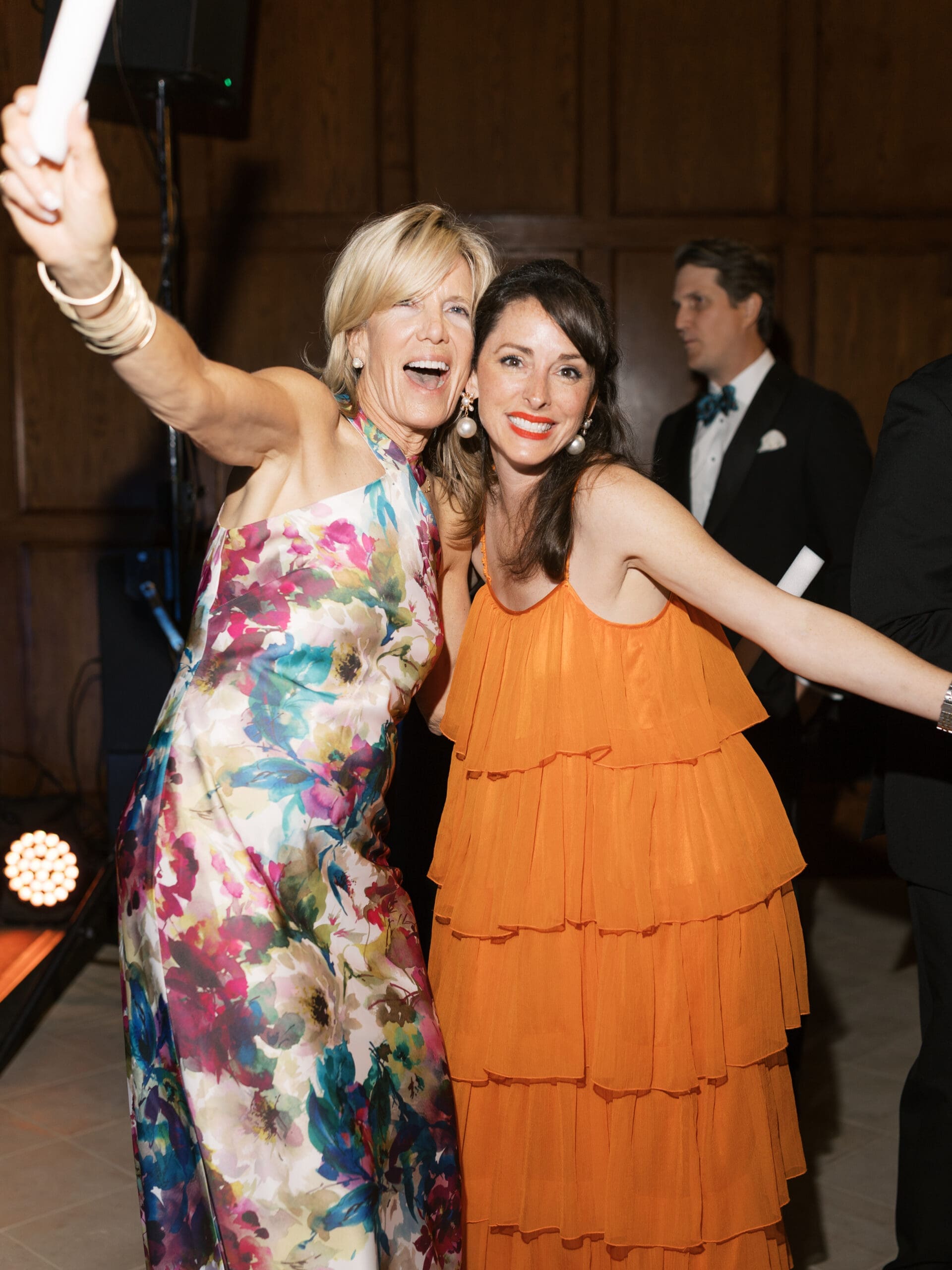 Two wedding guests in colorful gowns laughing and taking a selfie together on the reception dance floor.

