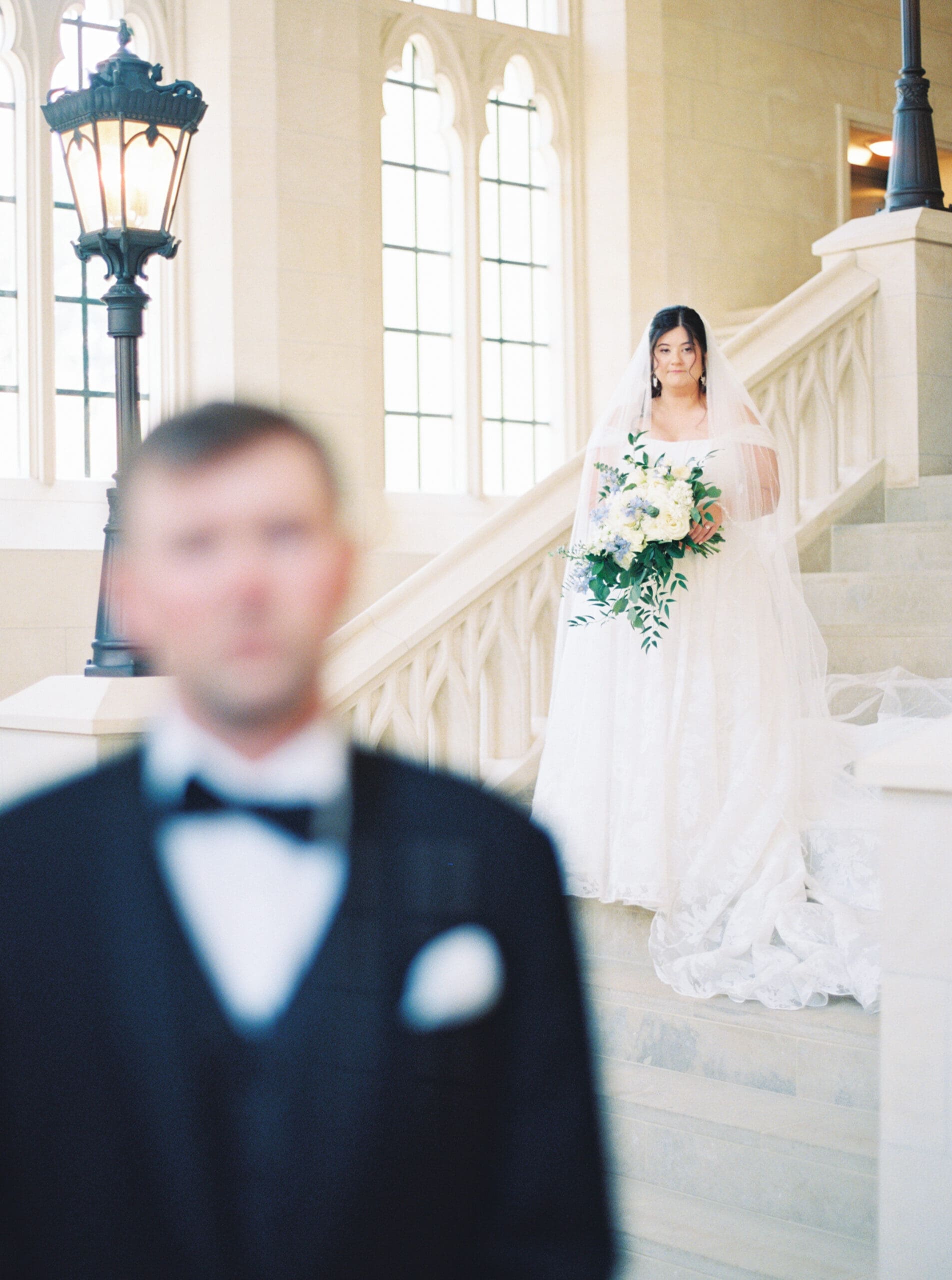 Bride standing on the staircase at The Lanier Theological Library holding her white and blue bouquet, with the groom blurred in the foreground.
