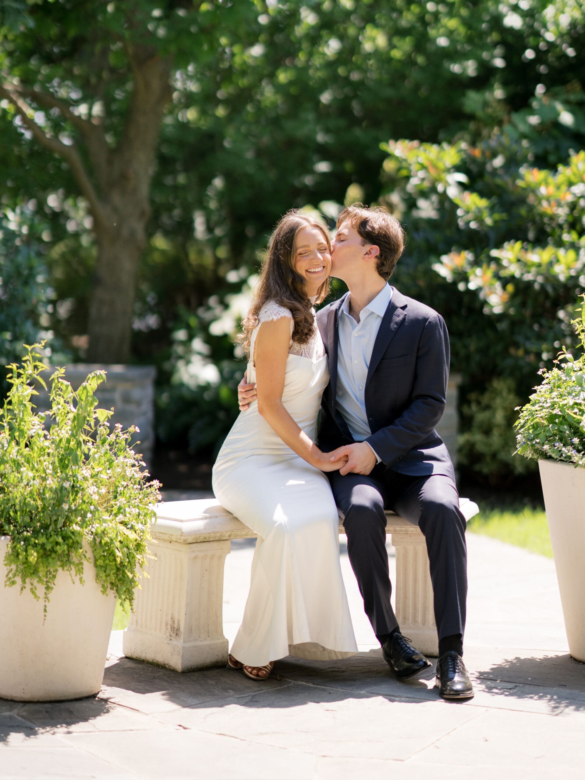The soon-to-be groom kissing his bride to be on the cheek while sitting together on a sunny garden bench.