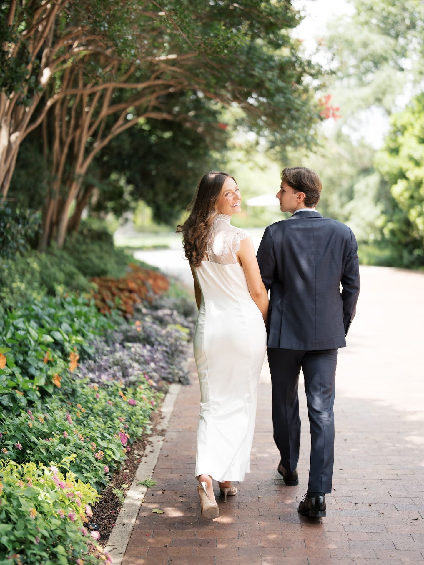 Couple strolling hand-in-hand down a brick pathway, glancing at each other affectionately.