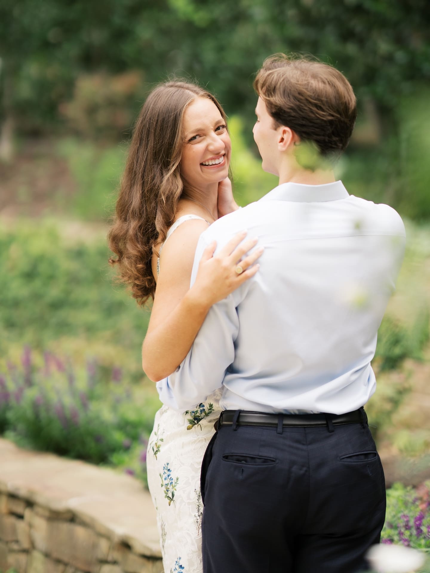 Bride-to-be glancing back with a joyful smile while wrapped in her fiancé's arms in a garden.
