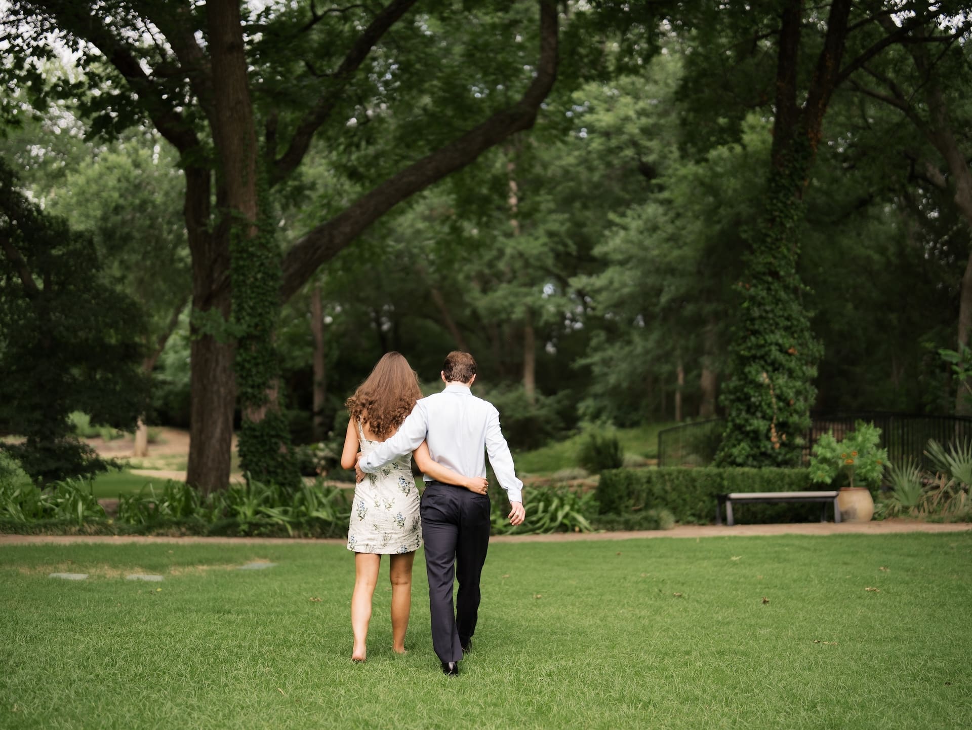Fall engagement photos of a couple walking away arm-in-arm beneath a canopy of trees.