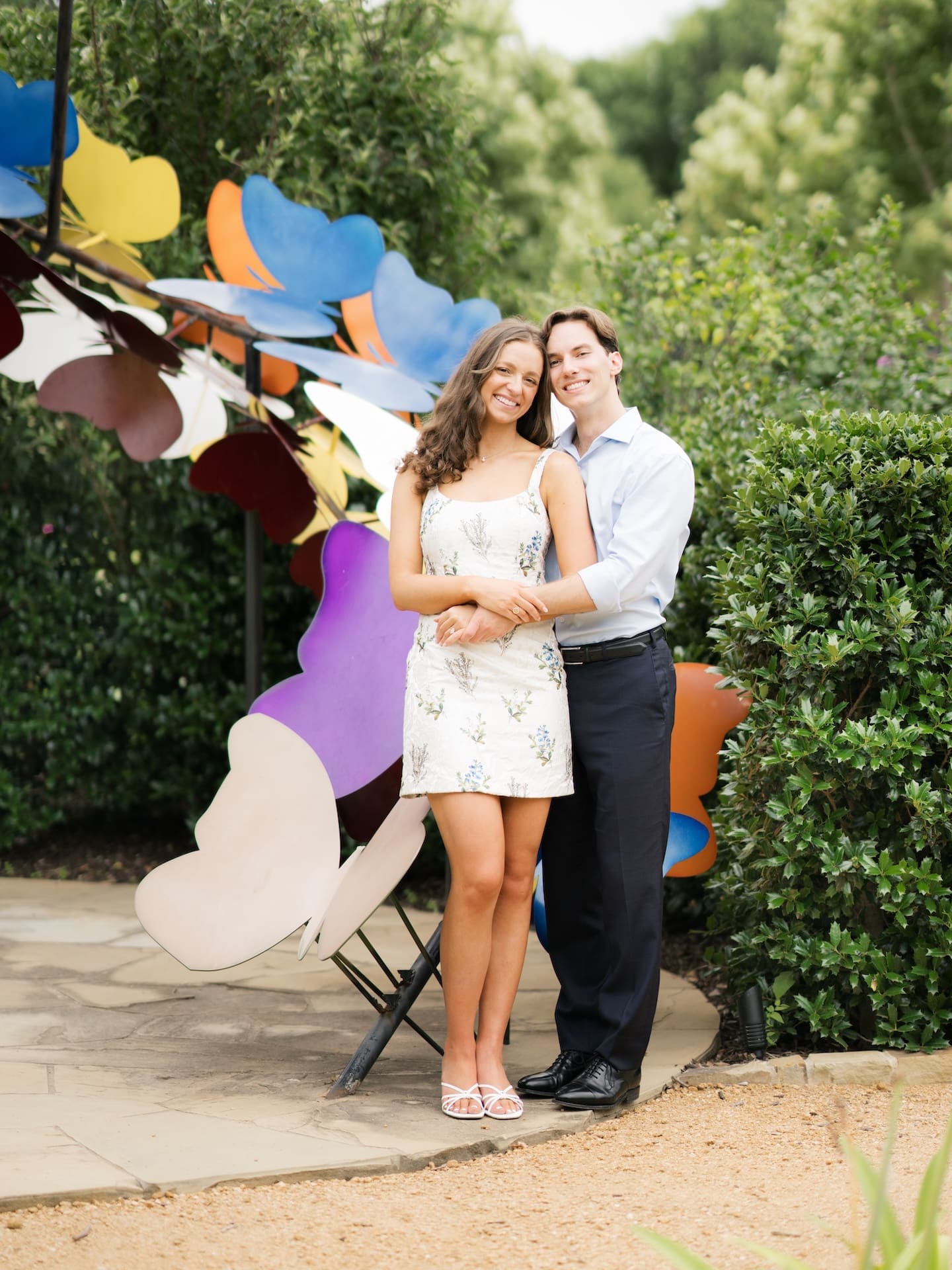 Couple smiling close together in front of a whimsical rainbow butterfly sculpture.
