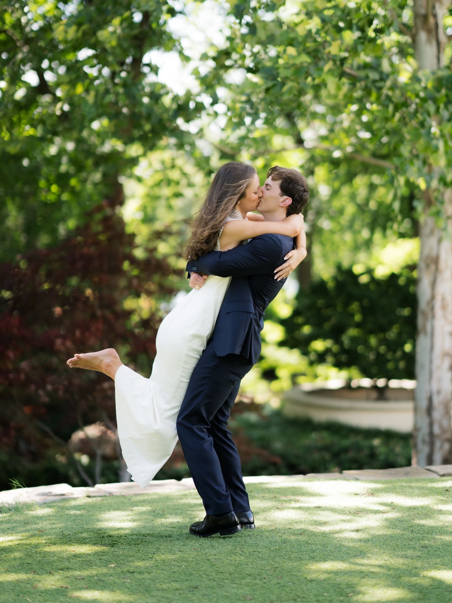 Groom lifting his bride-to-be off the ground as they share a kiss on the lawn.