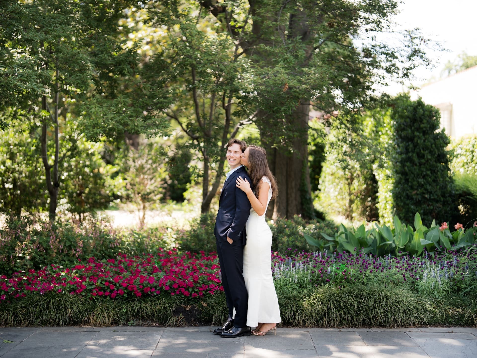 Bride-to-be kissing her fiancé's cheek as he grins beside a bright flower garden.